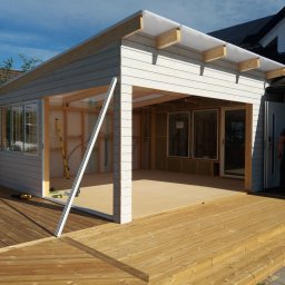 Krzysztof Kucharczyk - Drewniany taras połączony z budową szarej konstrukcji z oknami, widoczne narzędzia i fragment domu w tle, etap budowy.