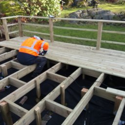 Krzysztof Kucharczyk - Montaż drewnianego tarasu: pracownik w odblaskowej kamizelce przykręca deski do ramy, widoczna konstrukcja nośna i częściowo ułożone deski podłogowe.