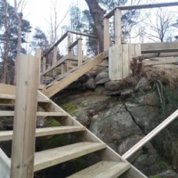 Drewniana konstrukcja schodów i balustrad w surowym stanie, wkomponowana w skalisty teren z drzewami w tle.