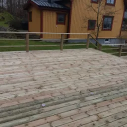 Nowa, jasna deska tarasowa z drewnianą balustradą na tle żółtego domu z czerwonymi obramowaniami okien.