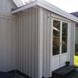 Krzysztof Kucharczyk - Zewnętrzna ściana budynku z drewnianą elewacją pomalowaną na jasny kolor, z widocznymi pionowymi deskami i oknem z drzwiami balkonowymi, obok którego stoi małe, czarne wiaderko.