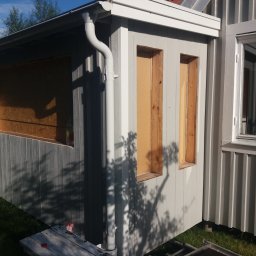 Krzysztof Kucharczyk - Fragment elewacji budynku z drewnianą konstrukcją okienną w trakcie montażu, widoczny system odprowadzania wody deszczowej, ściany pomalowane na szaro.