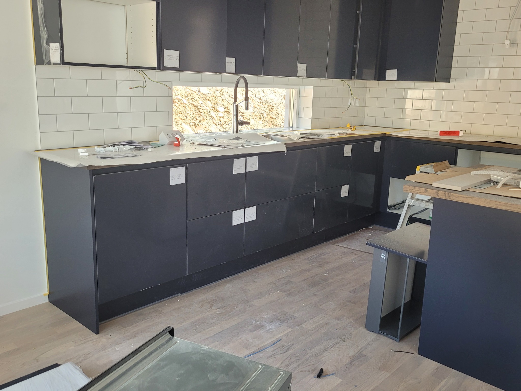 Montaż nowoczesnej, ciemnej zabudowy kuchennej w Szwecji, z widocznymi zabezpieczeniami na frontach szafek i białymi płytkami na ścianie, okno z widokiem na teren budowy, podłoga częściowo zakryta...