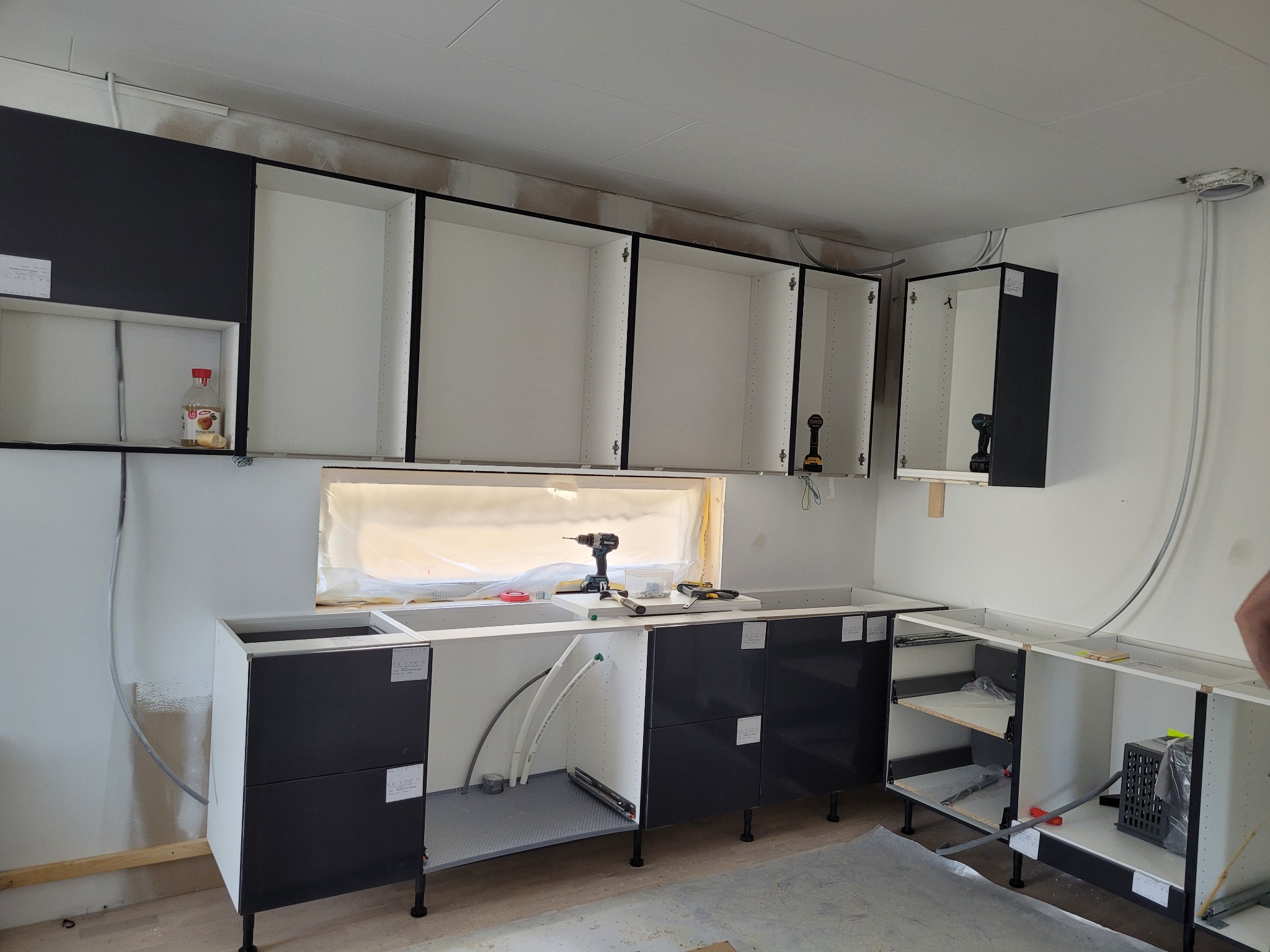 Montaż czarno-białych szafek kuchennych w surowym stanie, widoczne narzędzia i przewody, okno zasłonięte folią.