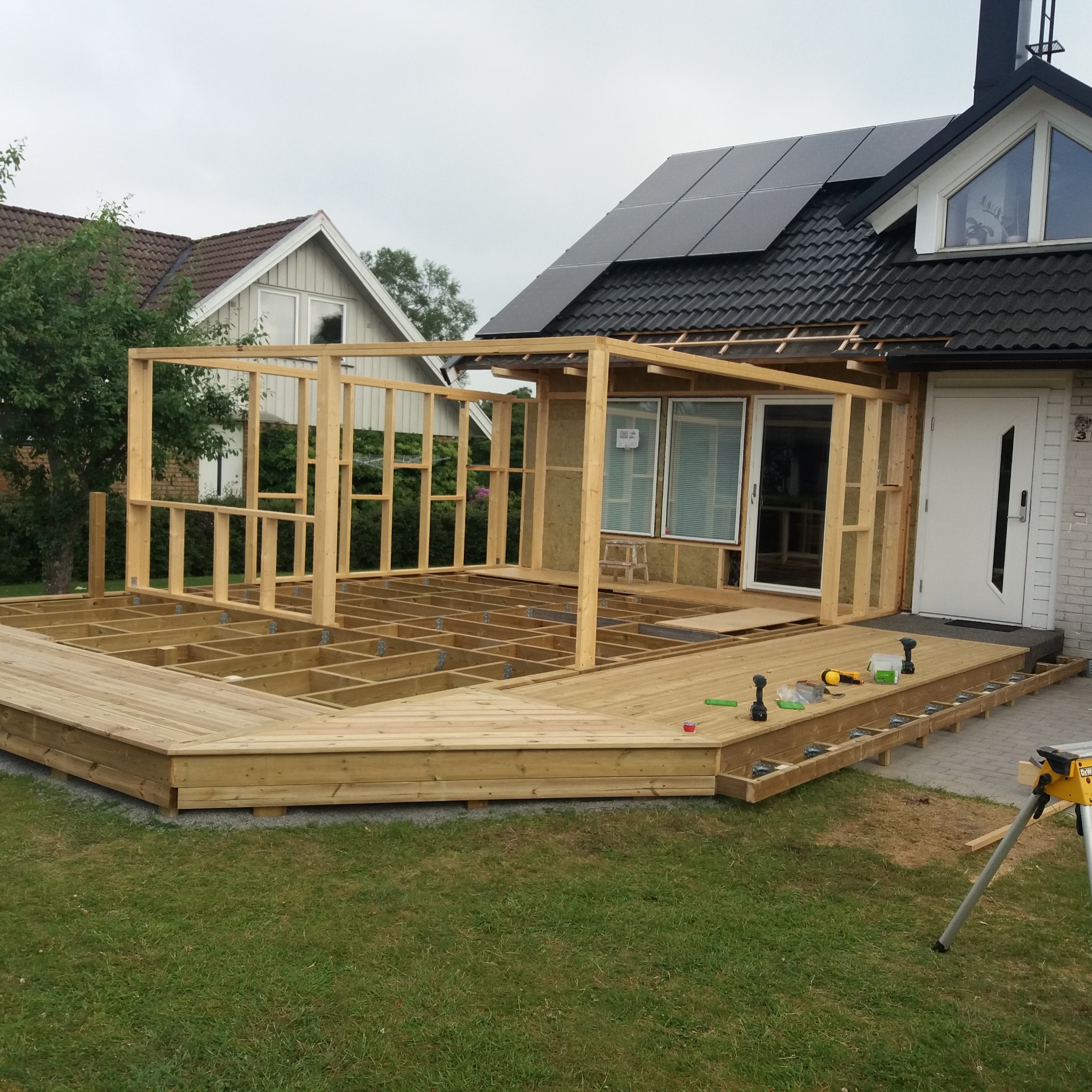 Budowa drewnianego tarasu przylegającego do domu z widoczną konstrukcją szkieletową z drewna, narzędziami i częściowo ułożonymi deskami podłogowymi.