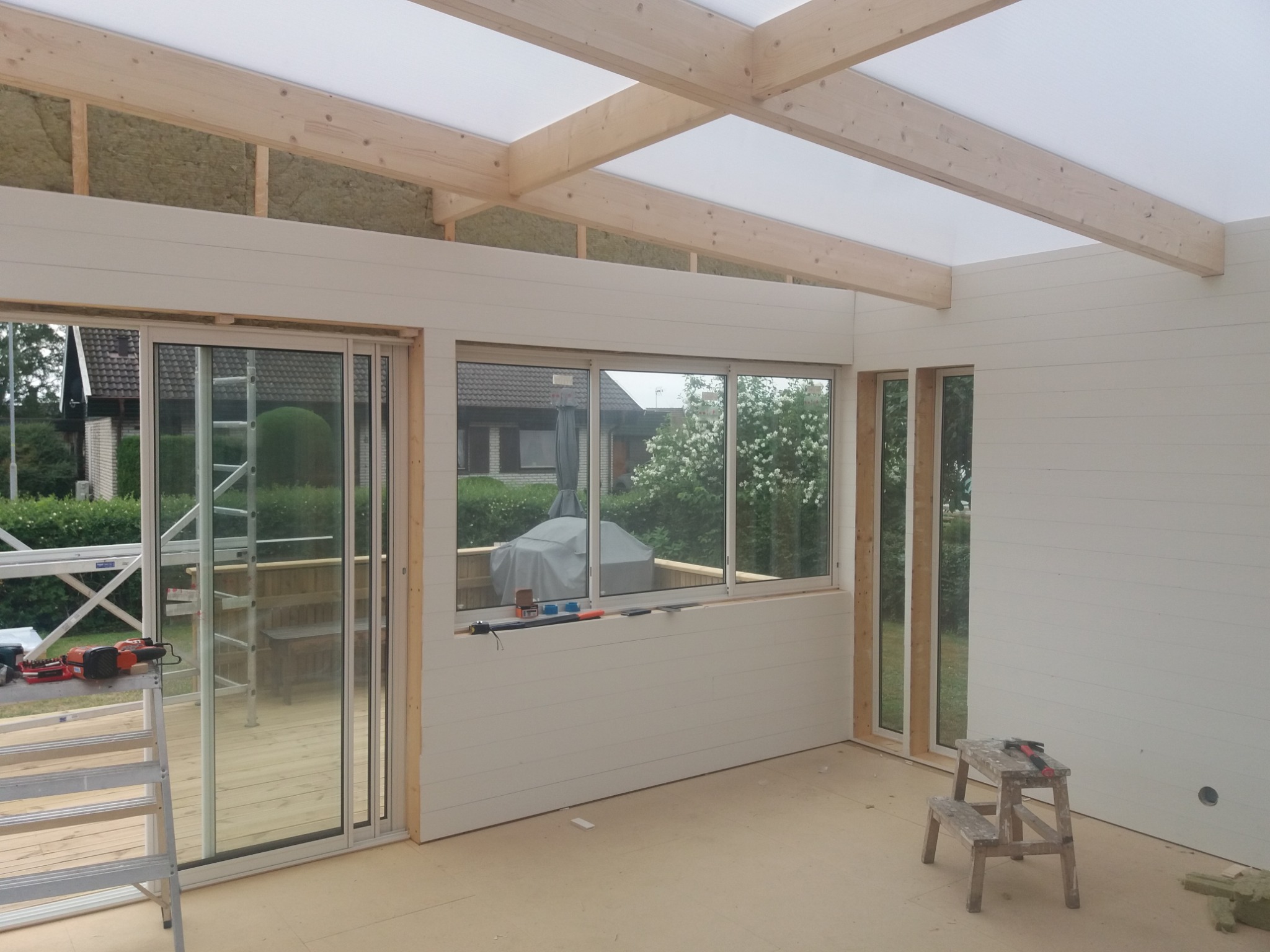 Wnętrze w trakcie budowy: surowe drewniane belki pod przezroczystym dachem, białe panele ścienne, okna z widokiem na zewnątrz, drabina i narzędzia sugerujące prace wykończeniowe.