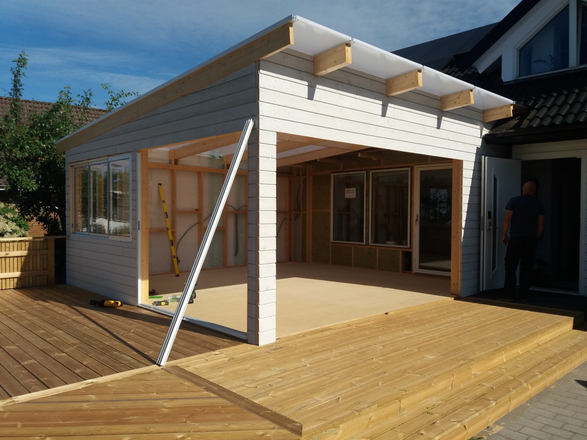 Drewniany taras połączony z budową szarej konstrukcji z oknami, widoczne narzędzia i fragment domu w tle, etap budowy.