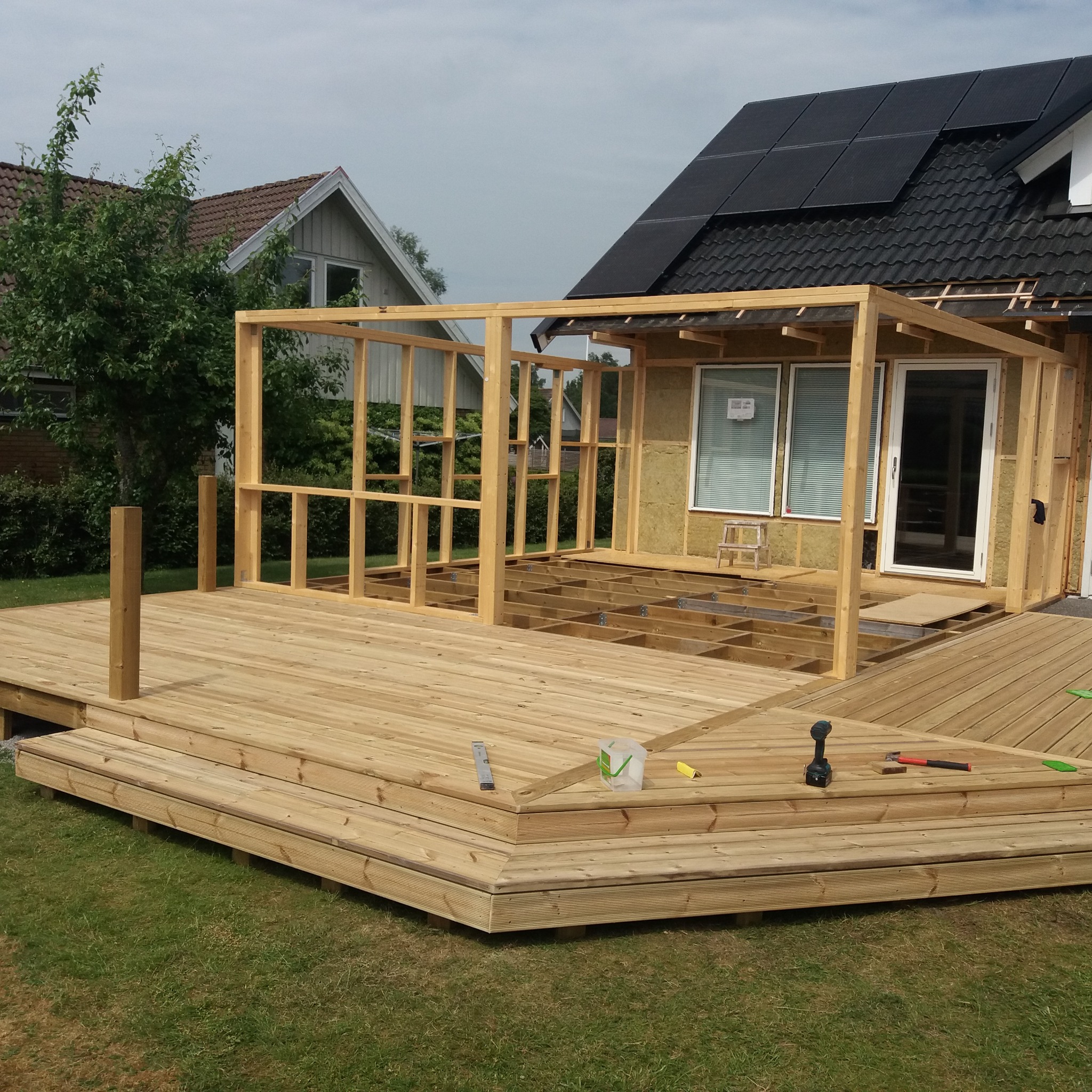 Budowa tarasu przylegającego do domu z panelami słonecznymi na dachu, widoczna konstrukcja drewniana i narzędzia pracy.