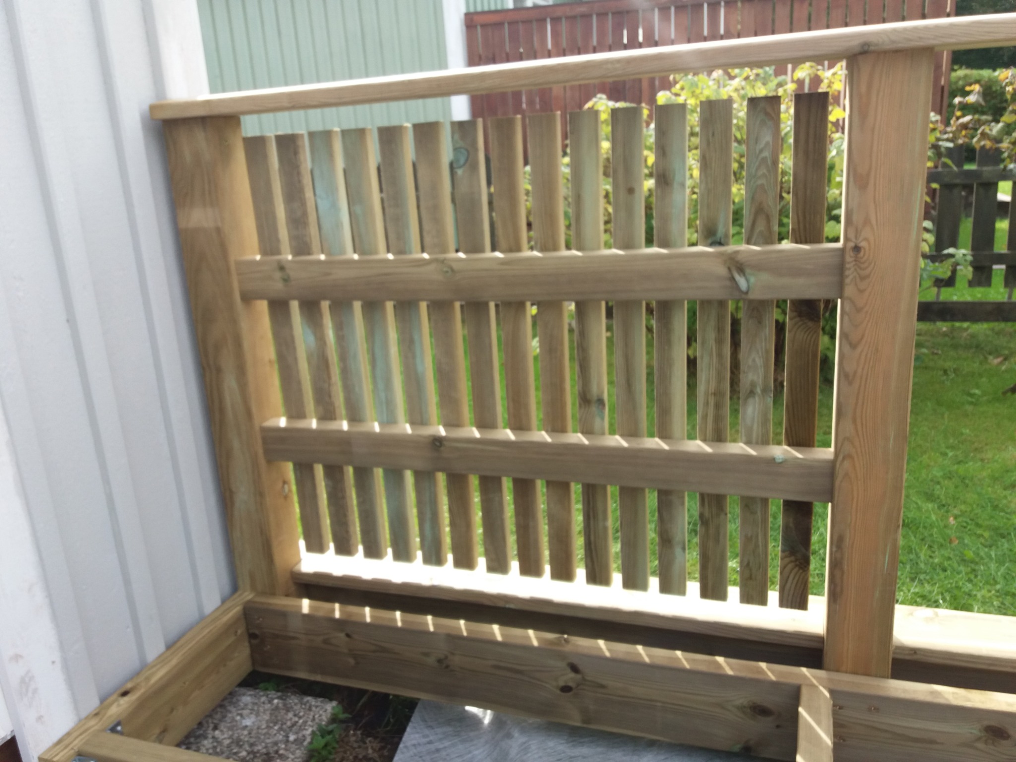 Fragment drewnianej konstrukcji tarasu z balustradą w trakcie budowy, widoczne elementy szkieletu i pionowe deski balustrady, tło stanowi trawnik i częściowo widoczne ogrodzenie.