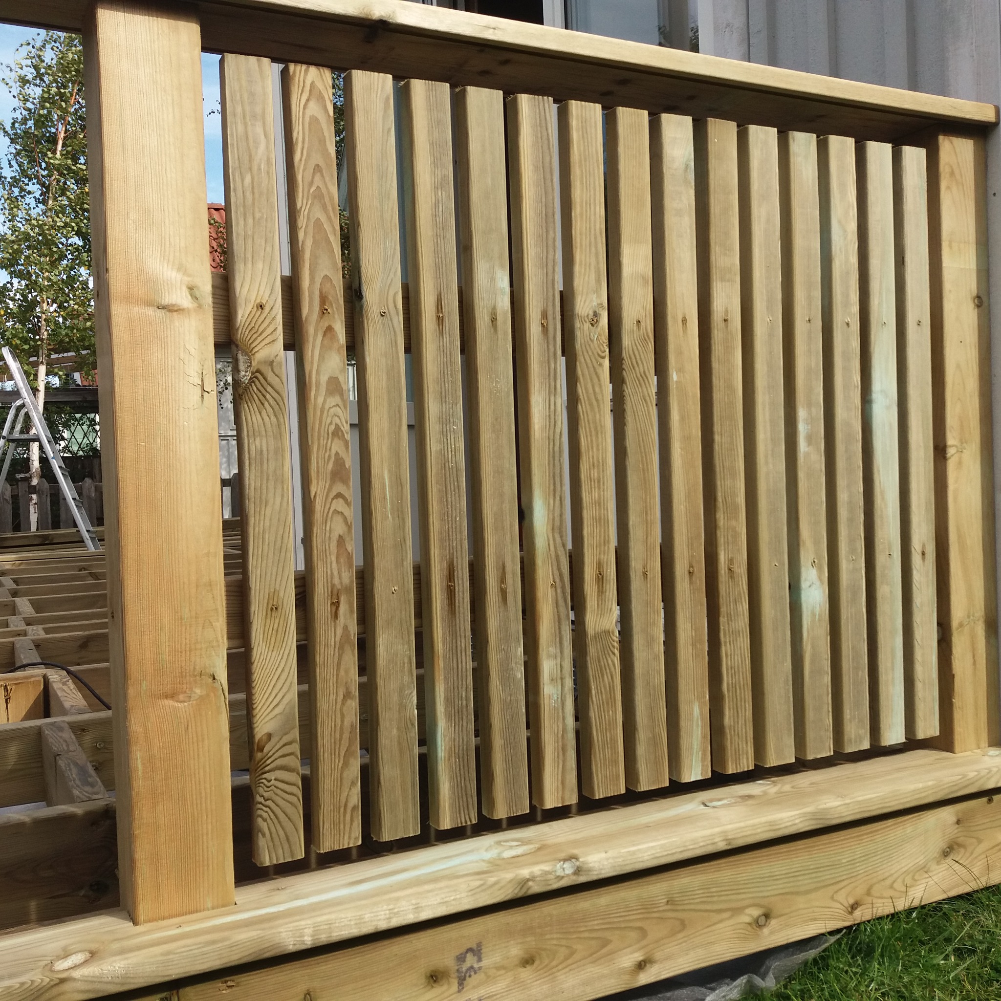 Fragment drewnianego tarasu w trakcie budowy, widoczna konstrukcja i pionowe deski balustrady, w tle drabina i zieleń.