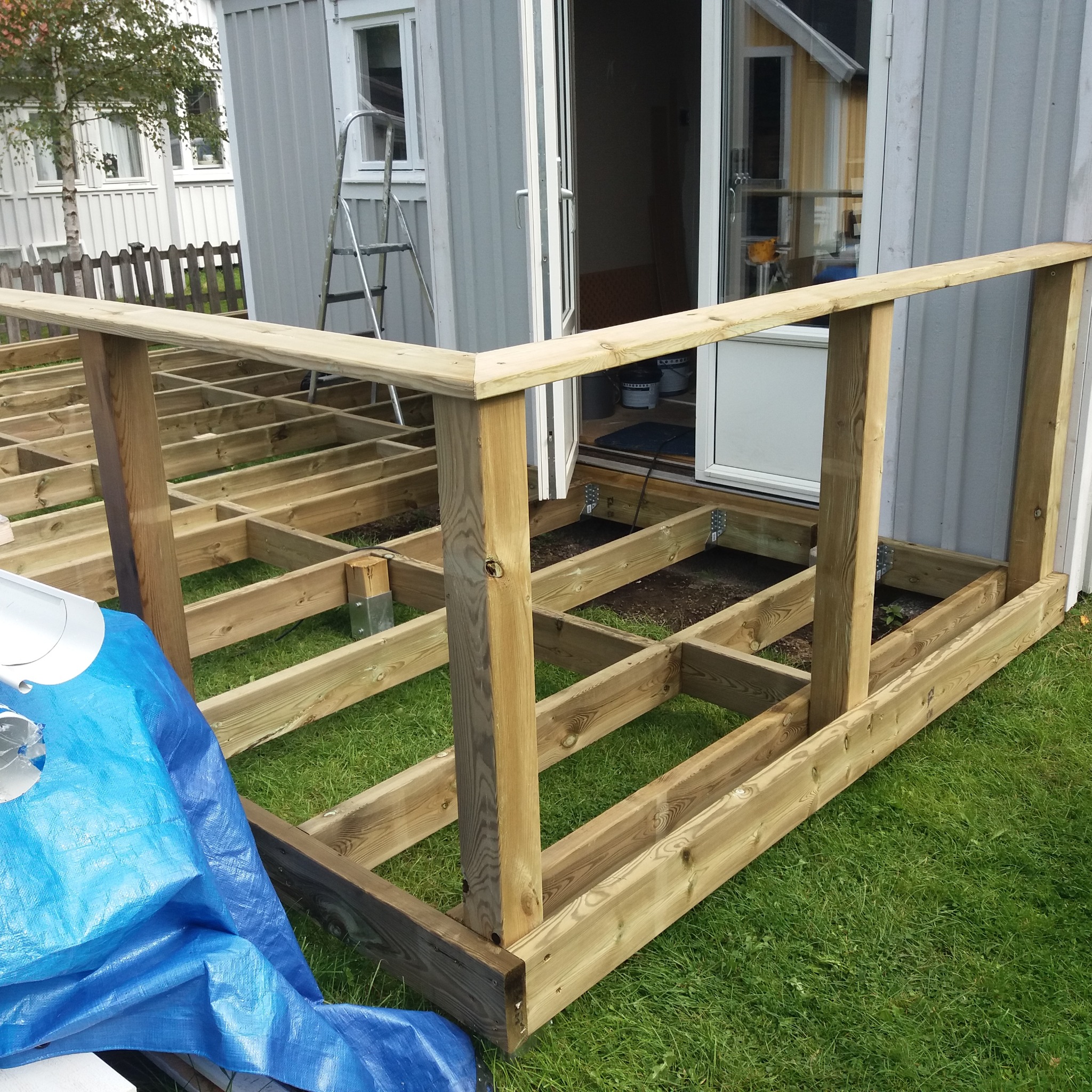Konstrukcja drewnianego tarasu w trakcie budowy, widoczne legary i słupy nośne, w tle dom z otwartymi drzwiami i drabina oparta o ścianę, na pierwszym planie niebieska plandeka.