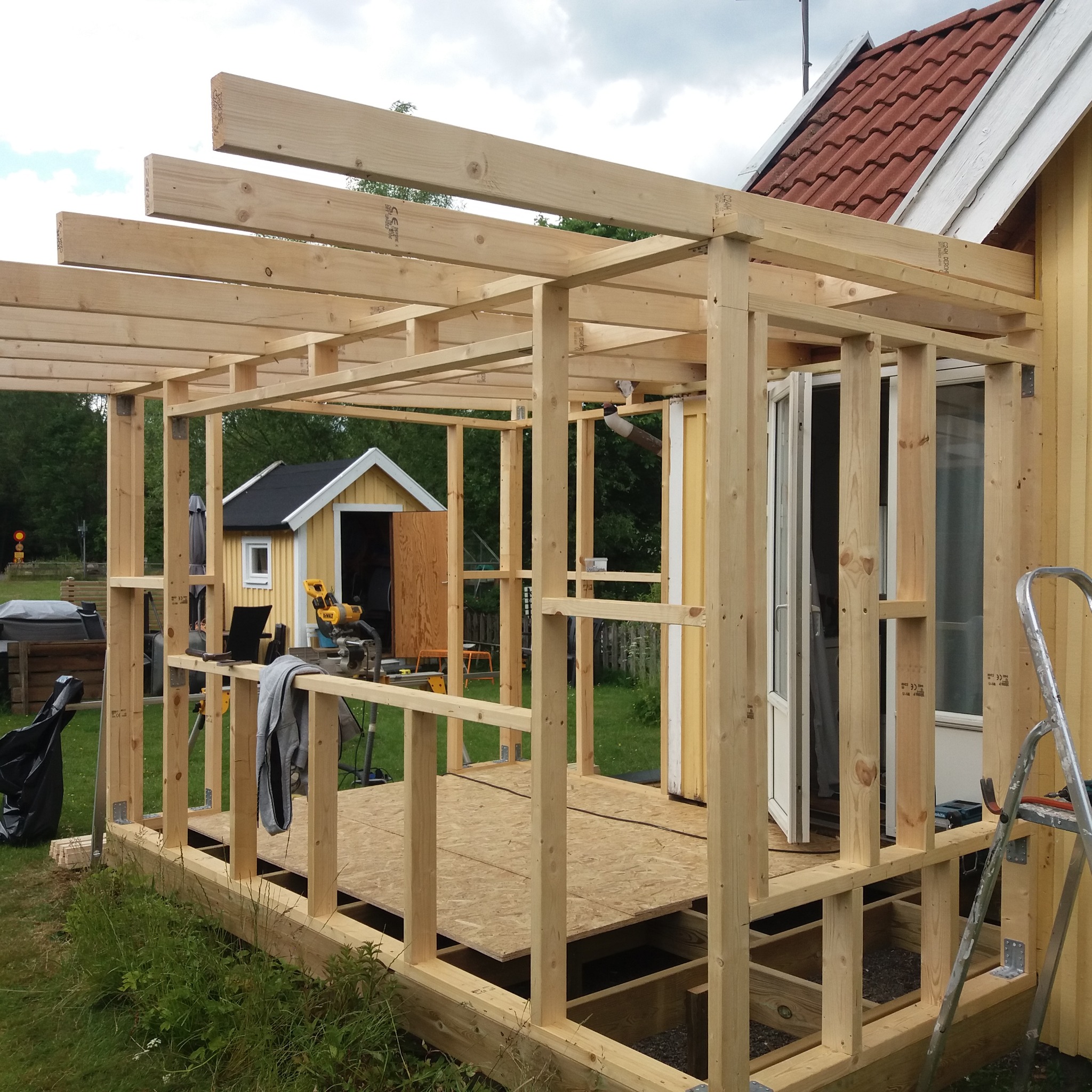 Konstrukcja szkieletowa drewnianego tarasu w trakcie budowy, z widocznymi elementami podłogi z płyt OSB, słupami nośnymi i belkami stropowymi; w tle żółty budynek z czerwonym dachem i narzędzia...