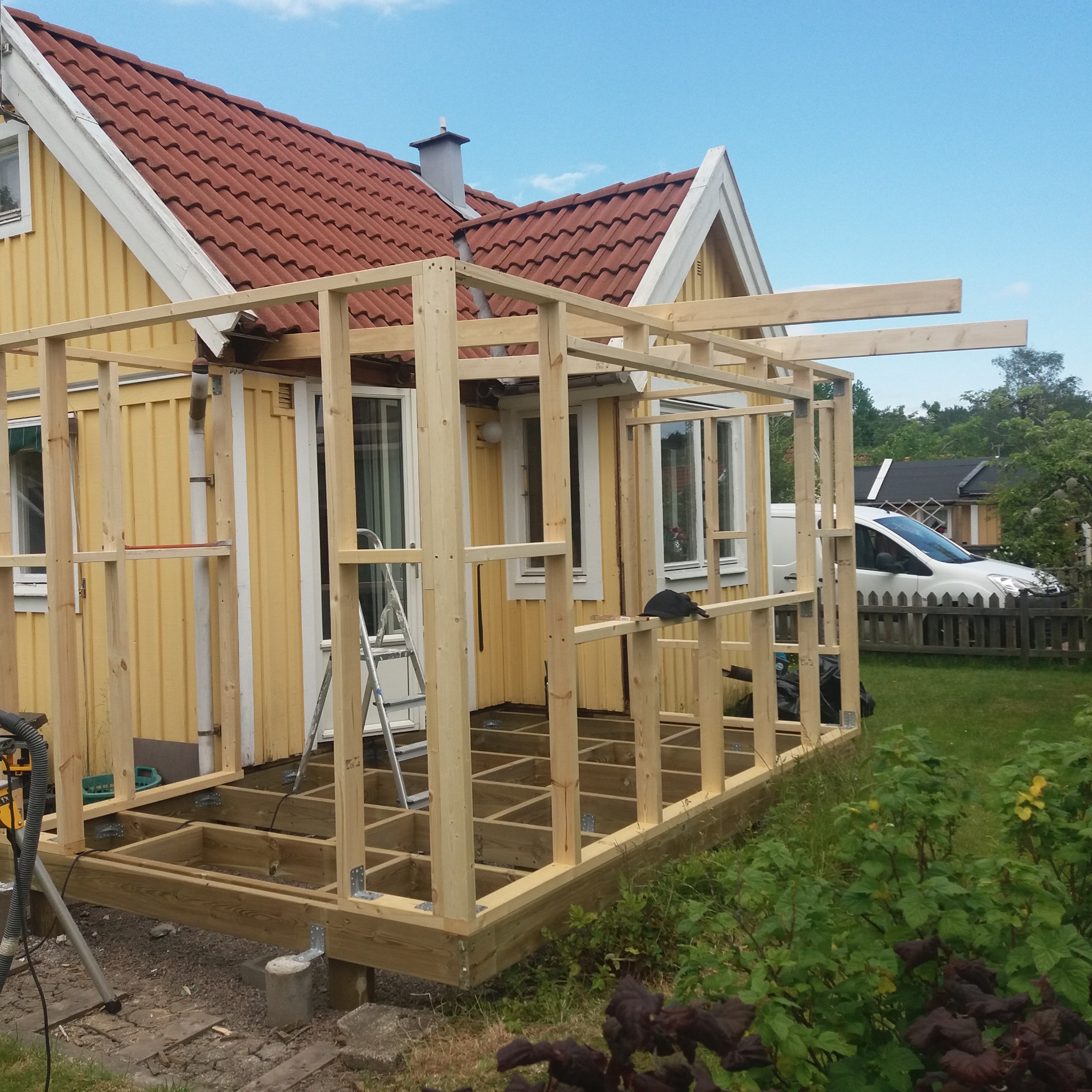 Konstrukcja szkieletowa z drewna przy żółtym domu z czerwonym dachem, widoczna drabina oparta o ścianę, częściowo zadaszony obszar, w tle biały samochód dostawczy.
