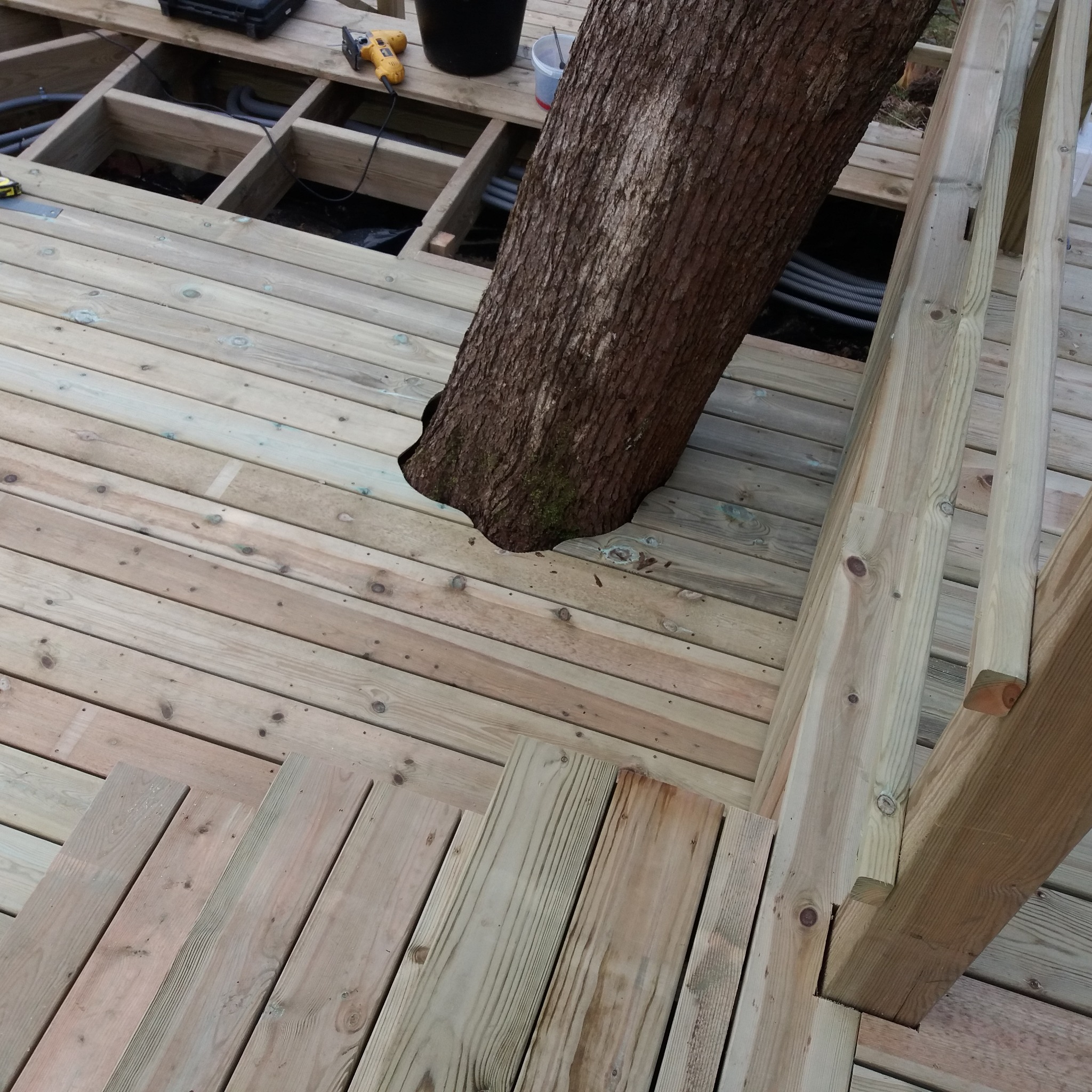 Konstrukcja drewnianego tarasu w trakcie budowy, z wkomponowanym pniem drzewa. Widoczne deski podłogowe, fragment schodów i balustrady oraz narzędzia pracy.