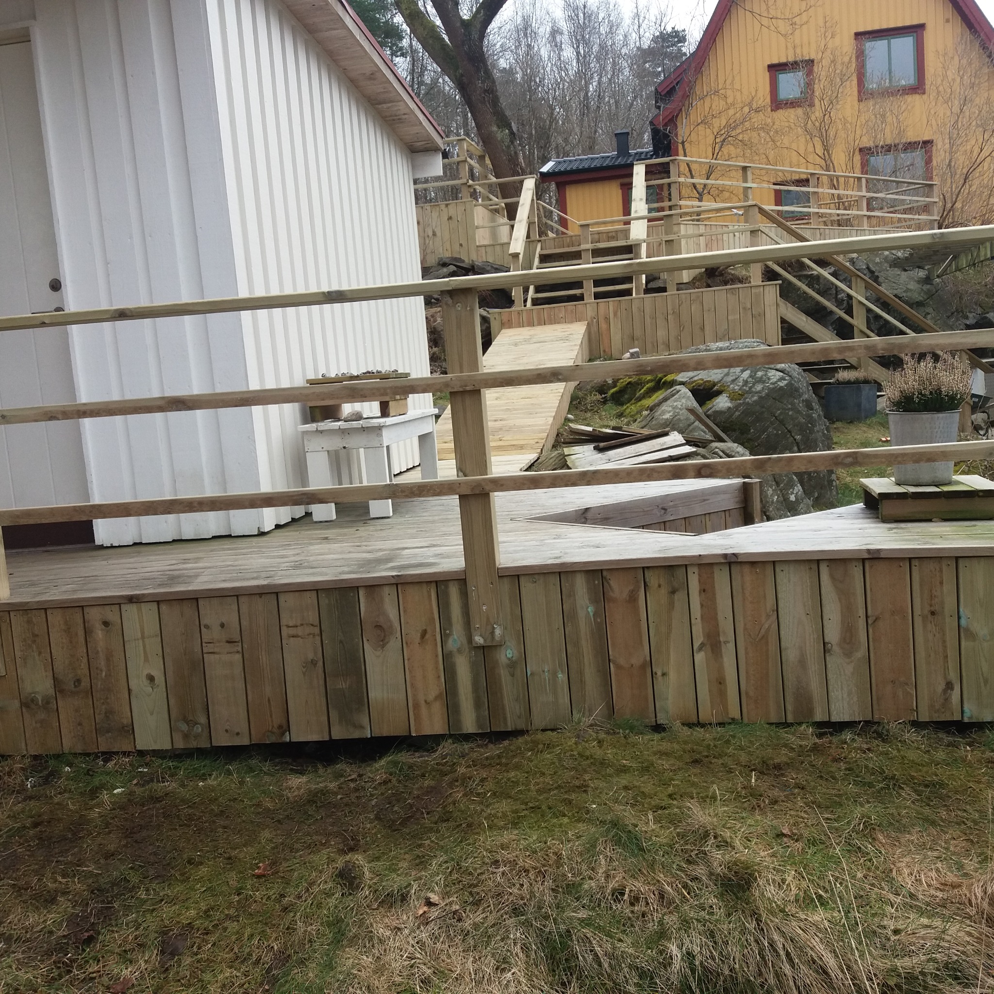 Wielopoziomowy taras z jasnego drewna z balustradami, łączący dwa budynki na nierównym terenie, z widocznymi schodami i rampą.