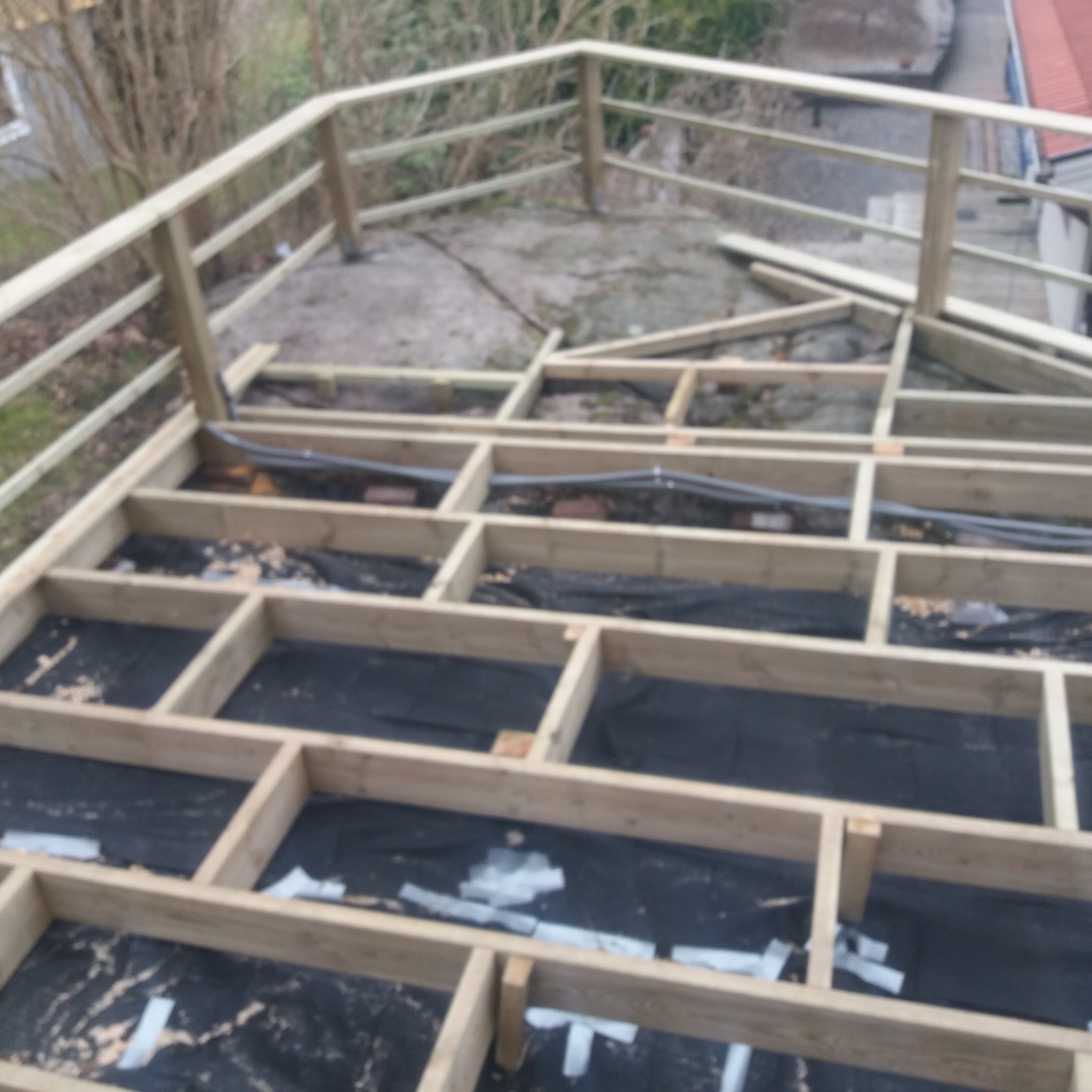 Konstrukcja drewniana tarasu w trakcie budowy, widoczne legary, folia izolacyjna i częściowo zamontowana balustrada z surowego drewna.