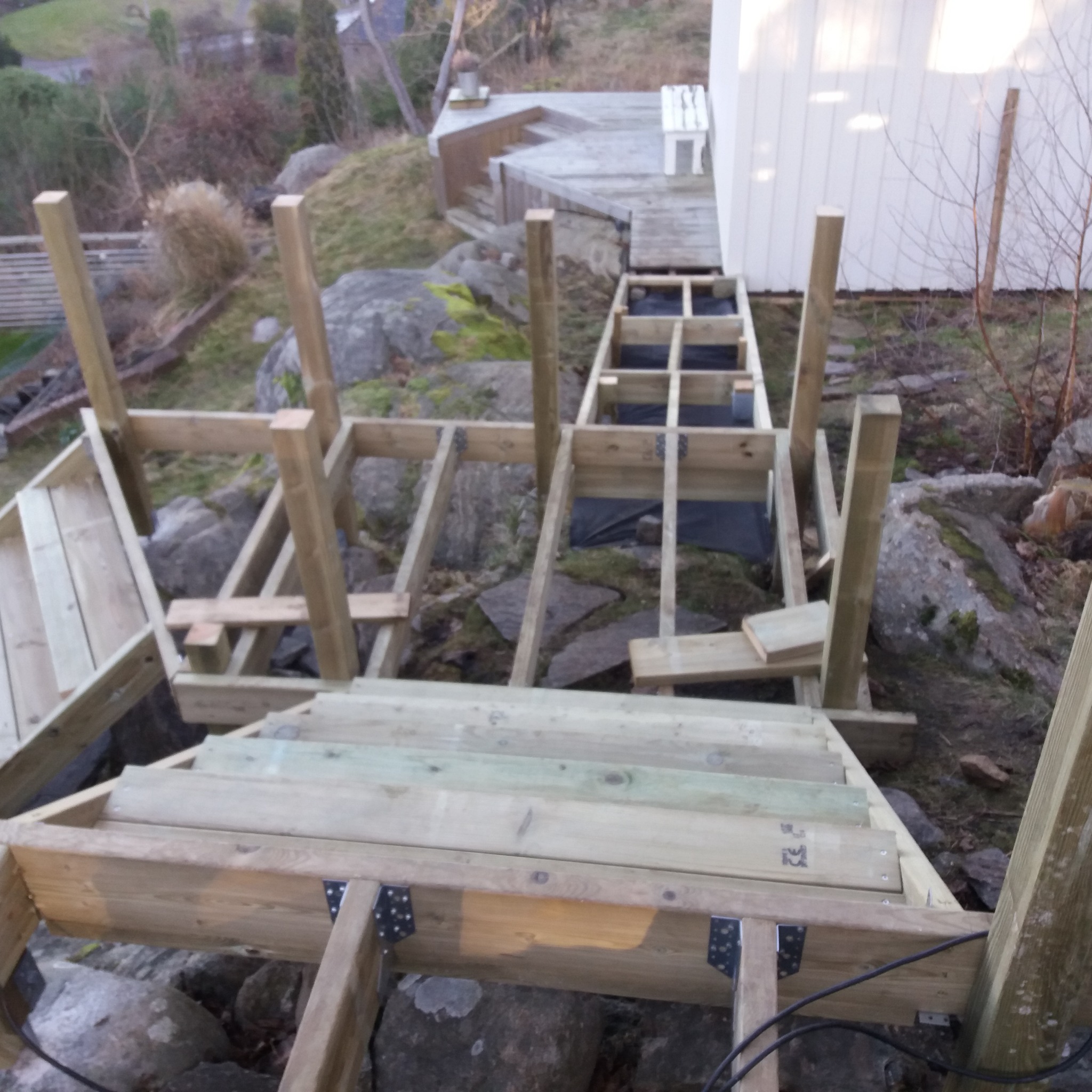 Konstrukcja drewnianego tarasu schodkowego w trakcie budowy na nierównym terenie skalnym, widoczne elementy nośne, schody z desek i czarna geowłóknina.