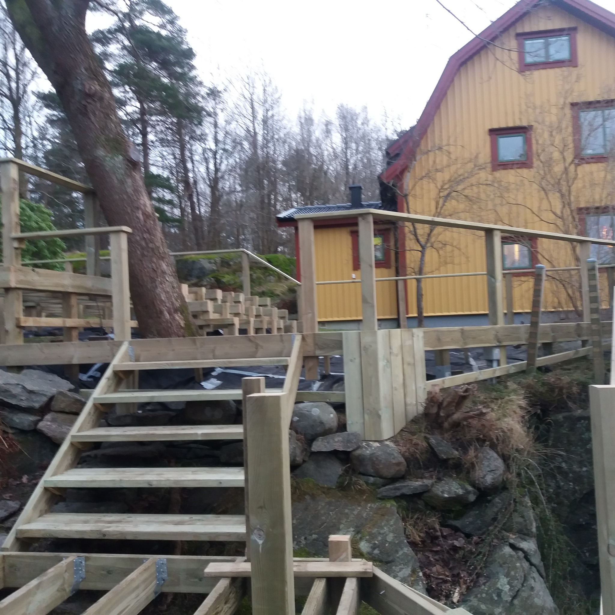 Konstrukcja drewnianego tarasu w trakcie budowy, z widocznymi schodami i balustradami, otoczona naturalnym skalistym terenem i drzewami, z żółtym domem w tle.