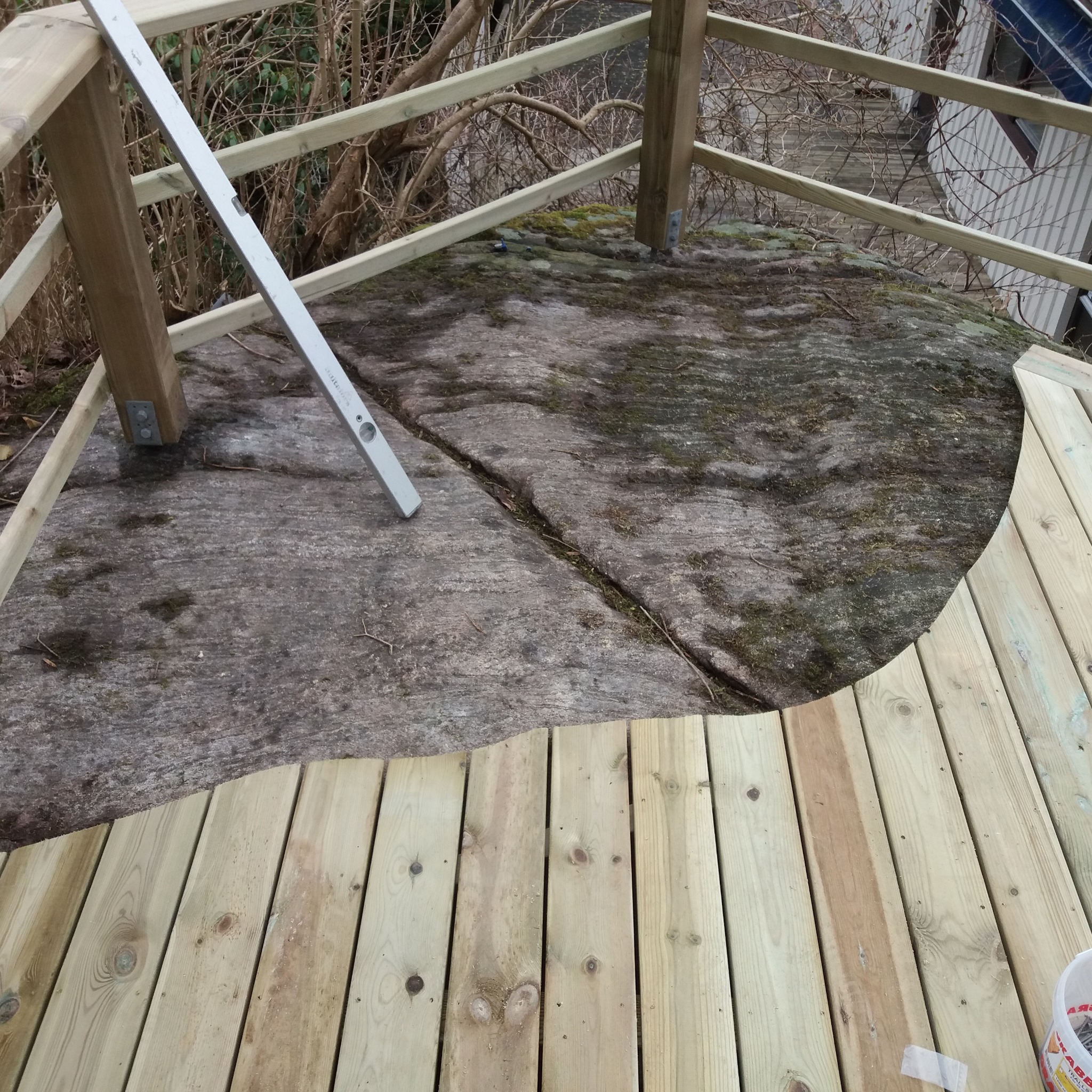 Drewniany taras w trakcie budowy na nierównym terenie skalnym; widoczna poziomica oparta o drewnianą konstrukcję balustrady, częściowo ułożone deski tarasowe z widocznymi słojami i sękami,...