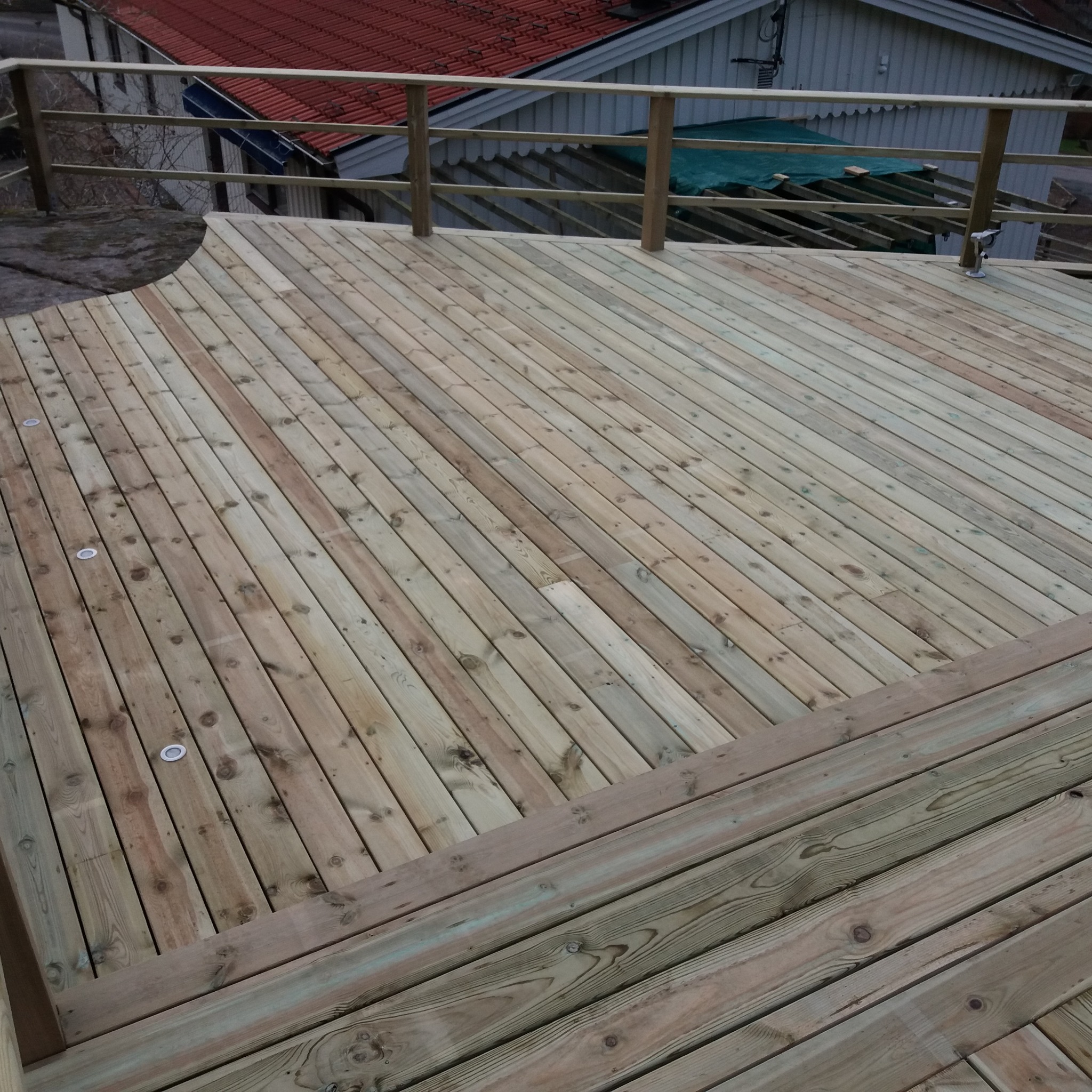 Nowo wybudowany taras z jasnego drewna z wbudowanymi punktowymi lampkami, otoczony drewnianą balustradą, w tle widoczny dom z czerwoną dachówką.