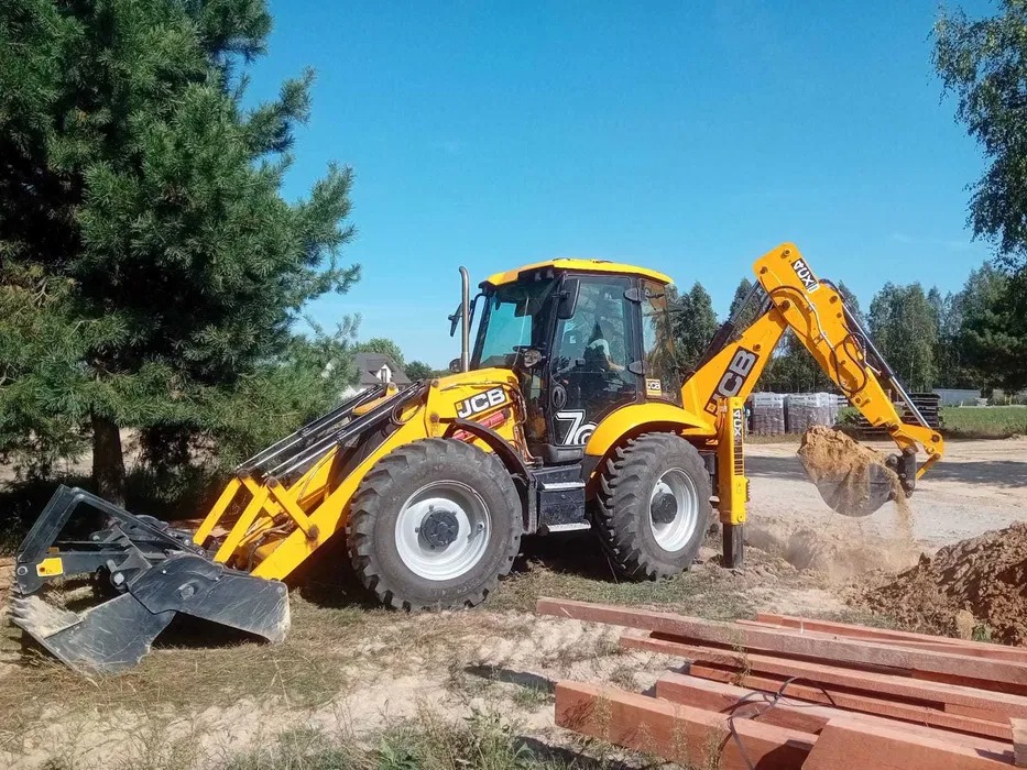 Żółta koparko-ładowarka JCB podczas wykopu ziemi, widoczne drewniane belki w tle, błękitne niebo i zielone drzewa.