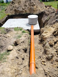 Wykopany dół z betonową płytą i kręgiem studziennym, do którego prowadzi pomarańczowa rura kanalizacyjna. Widoczny świeży nasyp ziemi i trawa w tle.