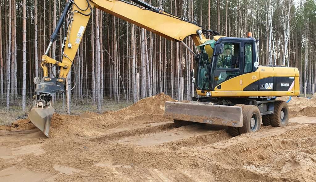 Żółta koparka kołowa CAT M316D z łyżką w trakcie prac ziemnych na piaszczystym terenie, w tle widoczny las sosnowy.