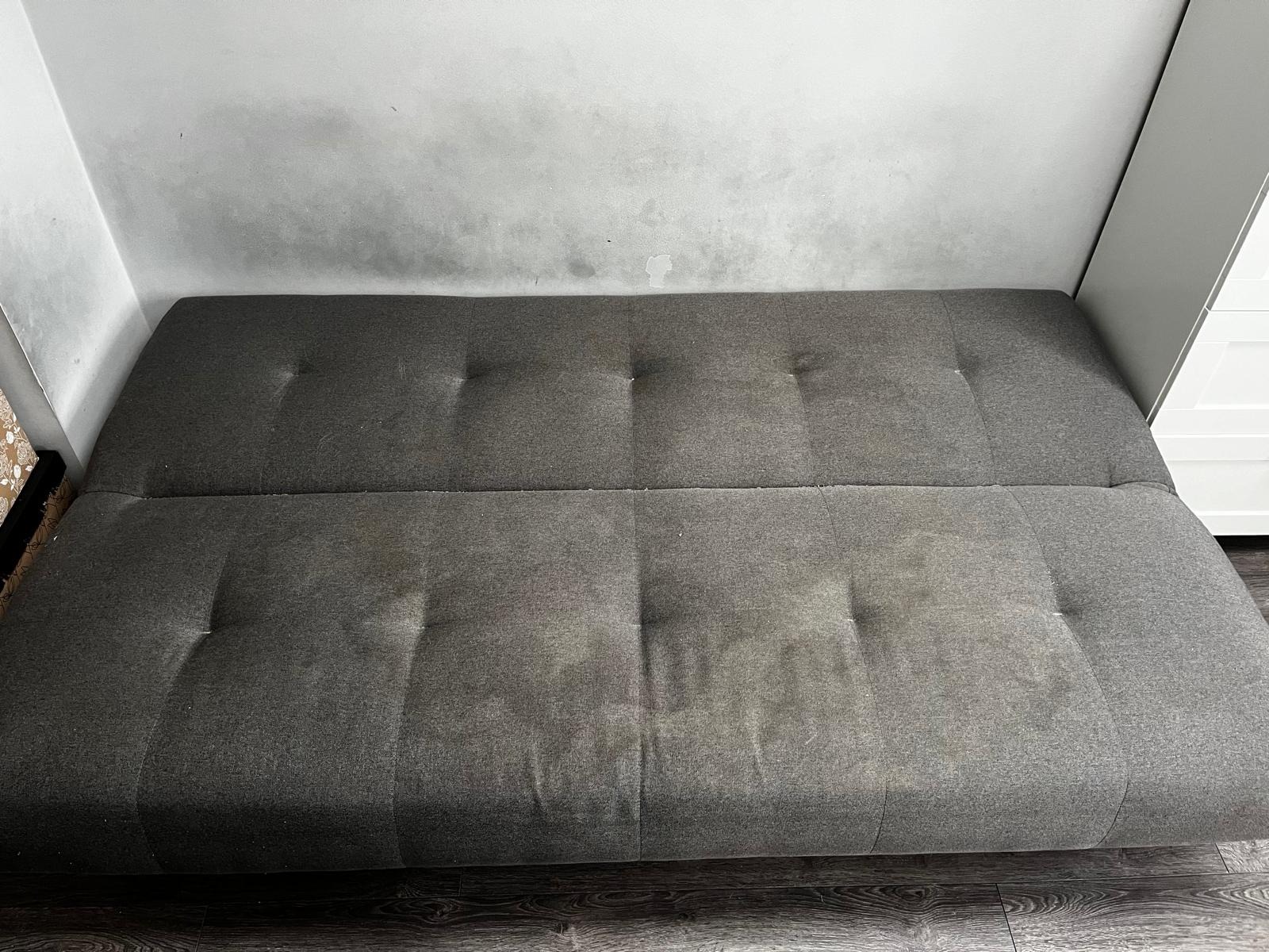 Szara, rozkładana kanapa z widocznymi plamami na siedzisku, ustawiona pod ścianą z zaciekami.