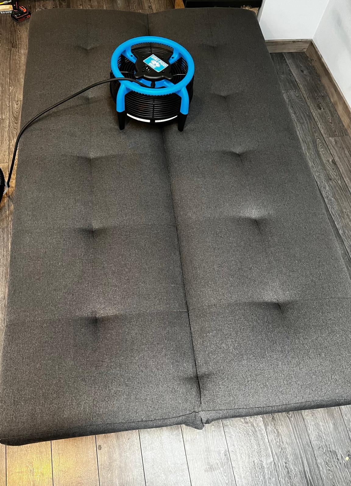 Szara, rozłożona sofa z tapicerowanymi guzikami, suszona niebiesko-czarnym wentylatorem do tapicerki.