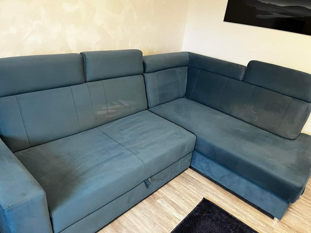 Rozłożona narożna kanapa z tapicerką w odcieniach szarości, widoczne ślady po czyszczeniu na siedzisku i oparciu, wysunięta szuflada na pościel.