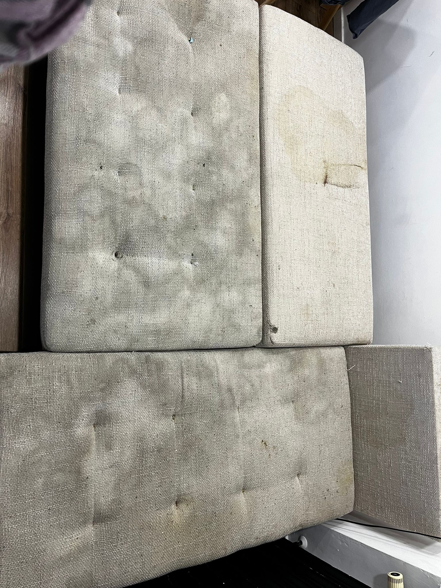 Fragment tapicerowanej kanapy z widocznymi plamami i zabrudzeniami na jasnej tkaninie obiciowej z guzikami.