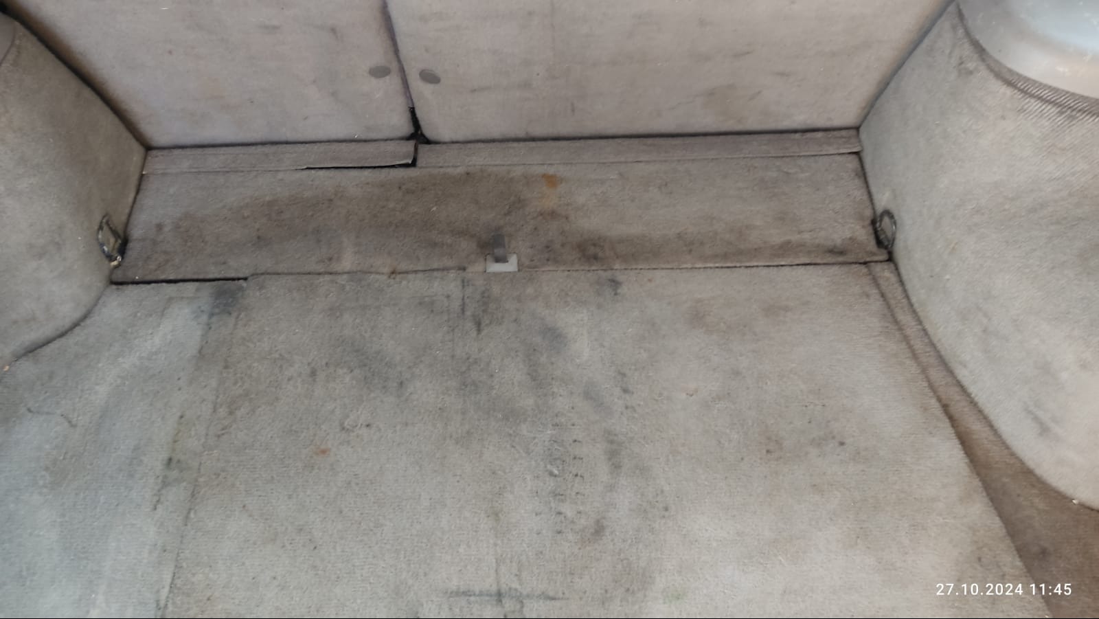 Wnętrze bagażnika samochodu z widocznymi plamami i zabrudzeniami na wykładzinie, z metalowym uchwytem po lewej stronie.