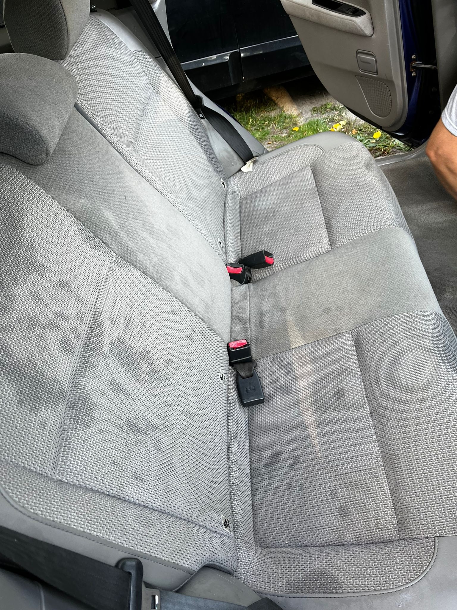 Tylna kanapa samochodu z szarej tapicerki z widocznymi plamami przed czyszczeniem, pasy bezpieczeństwa z czerwonymi elementami, otwarte drzwi auta i trawa z żółtymi kwiatami w tle.
