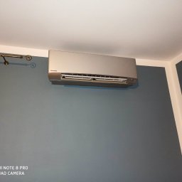 ENVERSO Bartosz Muzyka - Zamontowany klimatyzator Panasonic na szarej ścianie w pomieszczeniu, widoczna listwa karnisza i narożnik ściany z białą lamperią. Urządzenie w kolorze srebrnym.