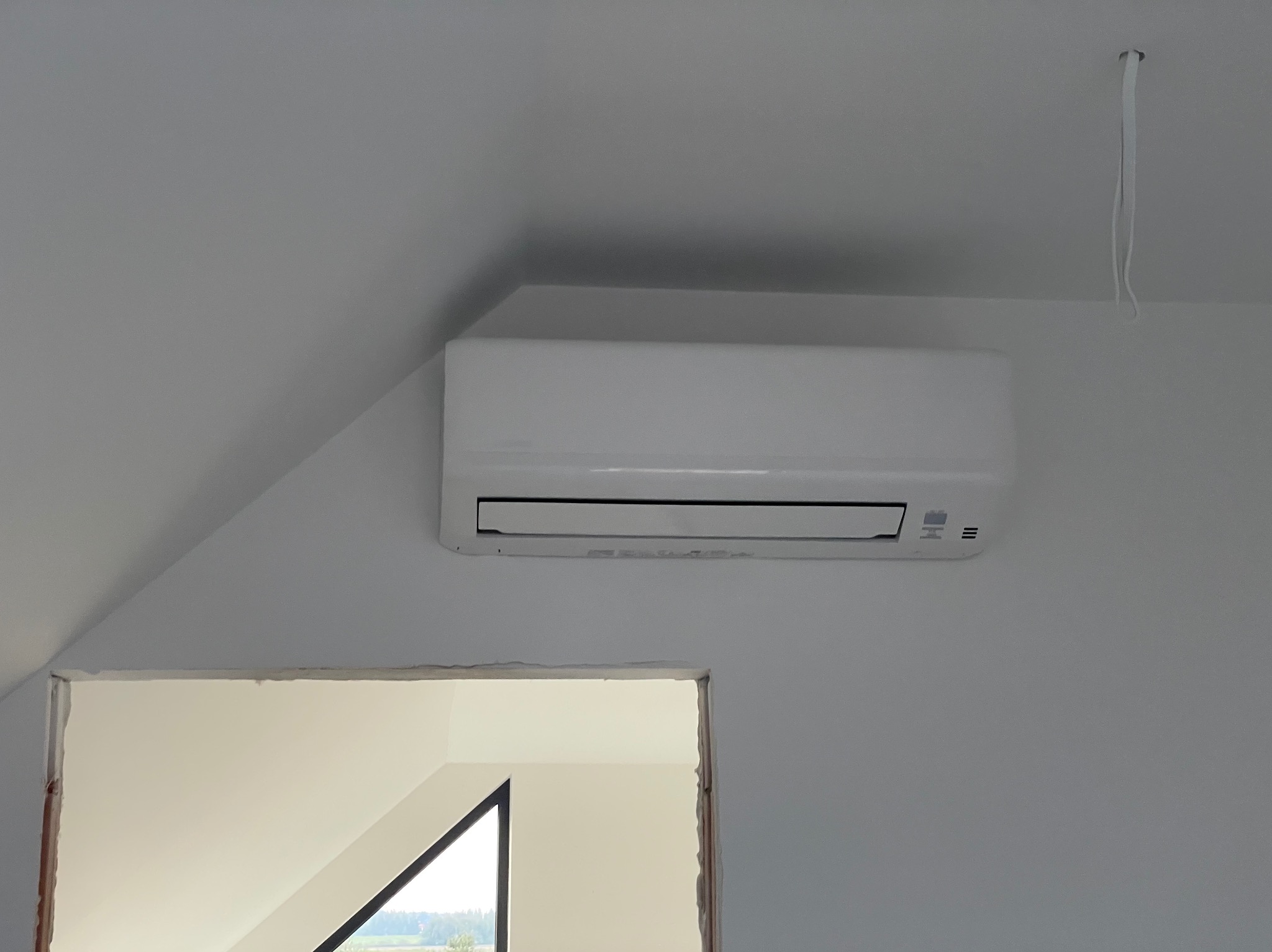 Biały klimatyzator ścienny zamontowany w jasnym pomieszczeniu, widoczny fragment niezamaskowanego otworu w ścianie i zwisający przewód.
