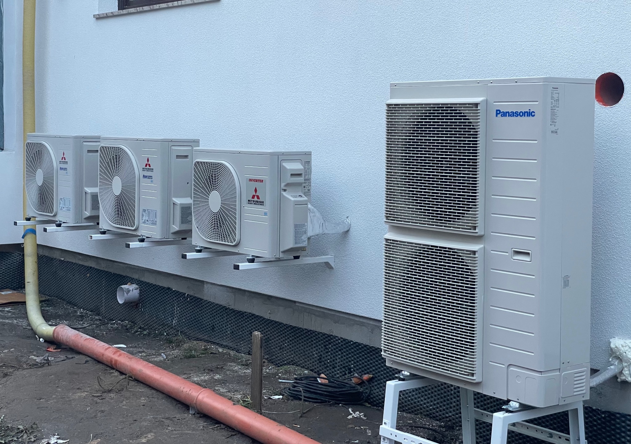 Zewnętrzne jednostki klimatyzacji Mitsubishi i Panasonic zamontowane na elewacji budynku, widoczne rury odprowadzające skropliny i przewody elektryczne, teren wokół w trakcie prac instalacyjnych.