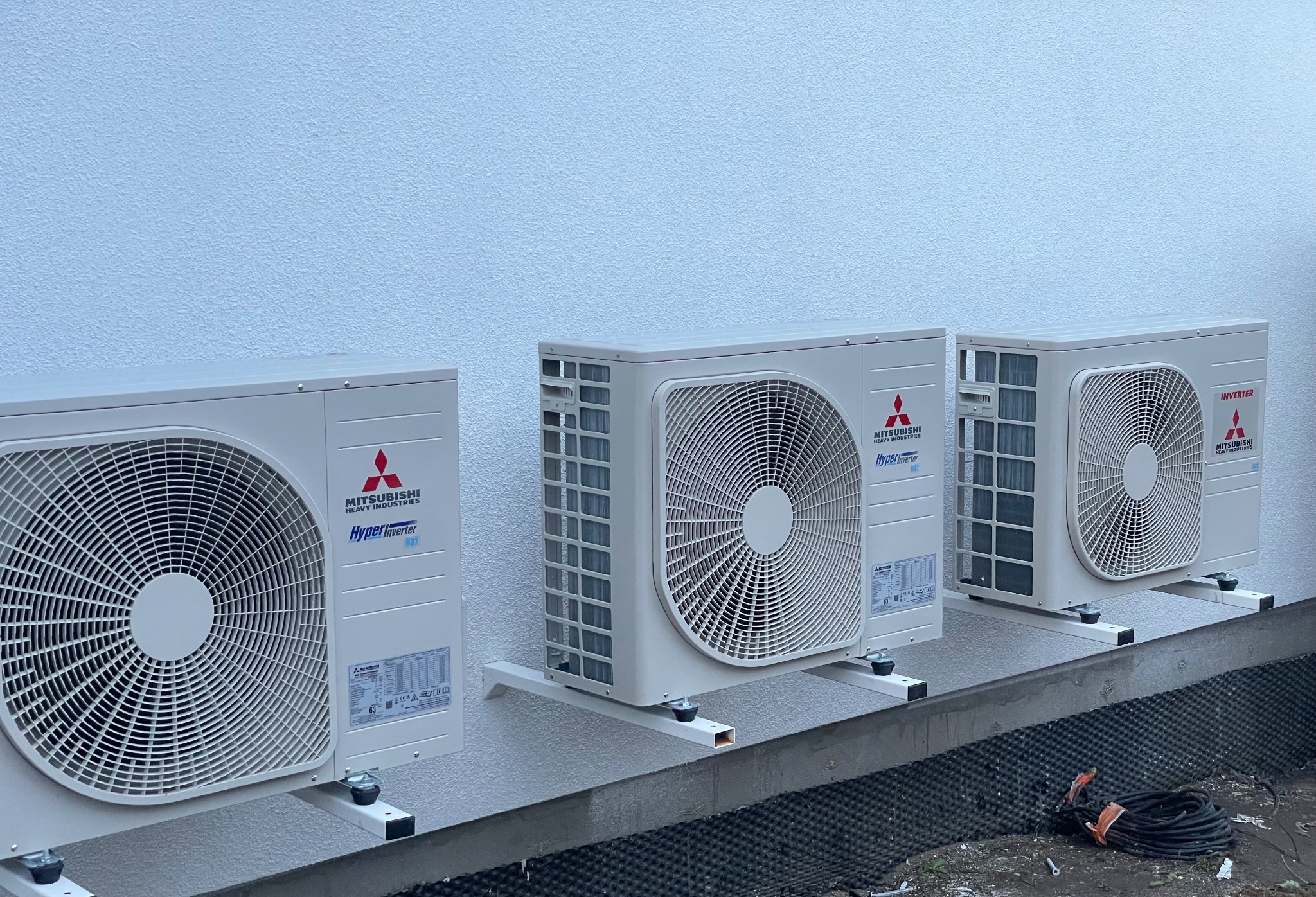 Trzy zewnętrzne jednostki klimatyzacyjne Mitsubishi Hyper Inverter zamontowane na białej ścianie budynku, z widocznymi wspornikami i częścią instalacji elektrycznej na ziemi.
