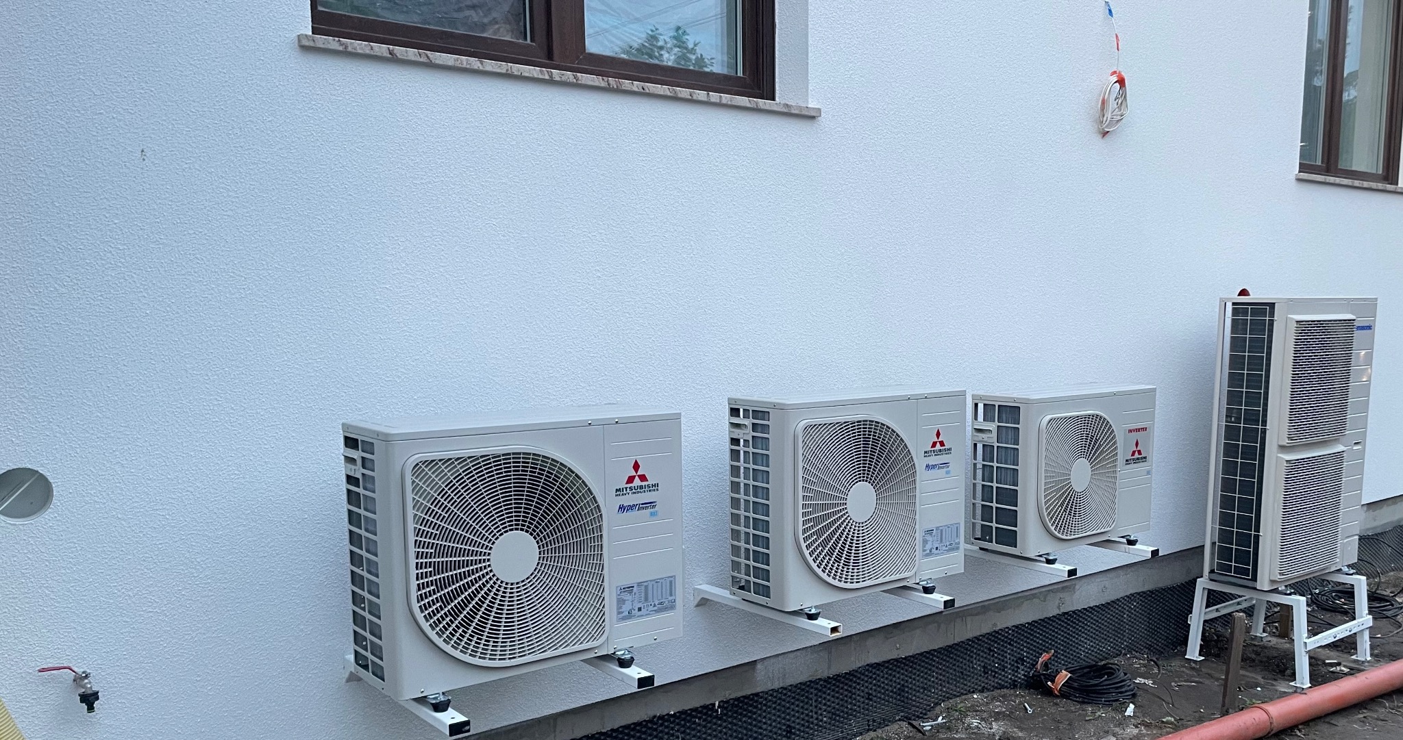 Trzy jednostki zewnętrzne klimatyzacji Mitsubishi Hyper Inverter zamontowane na wspornikach przy białej, teksturowanej ścianie budynku, z widoczną drugą jednostką zewnętrzną Panasonic na metalowej...