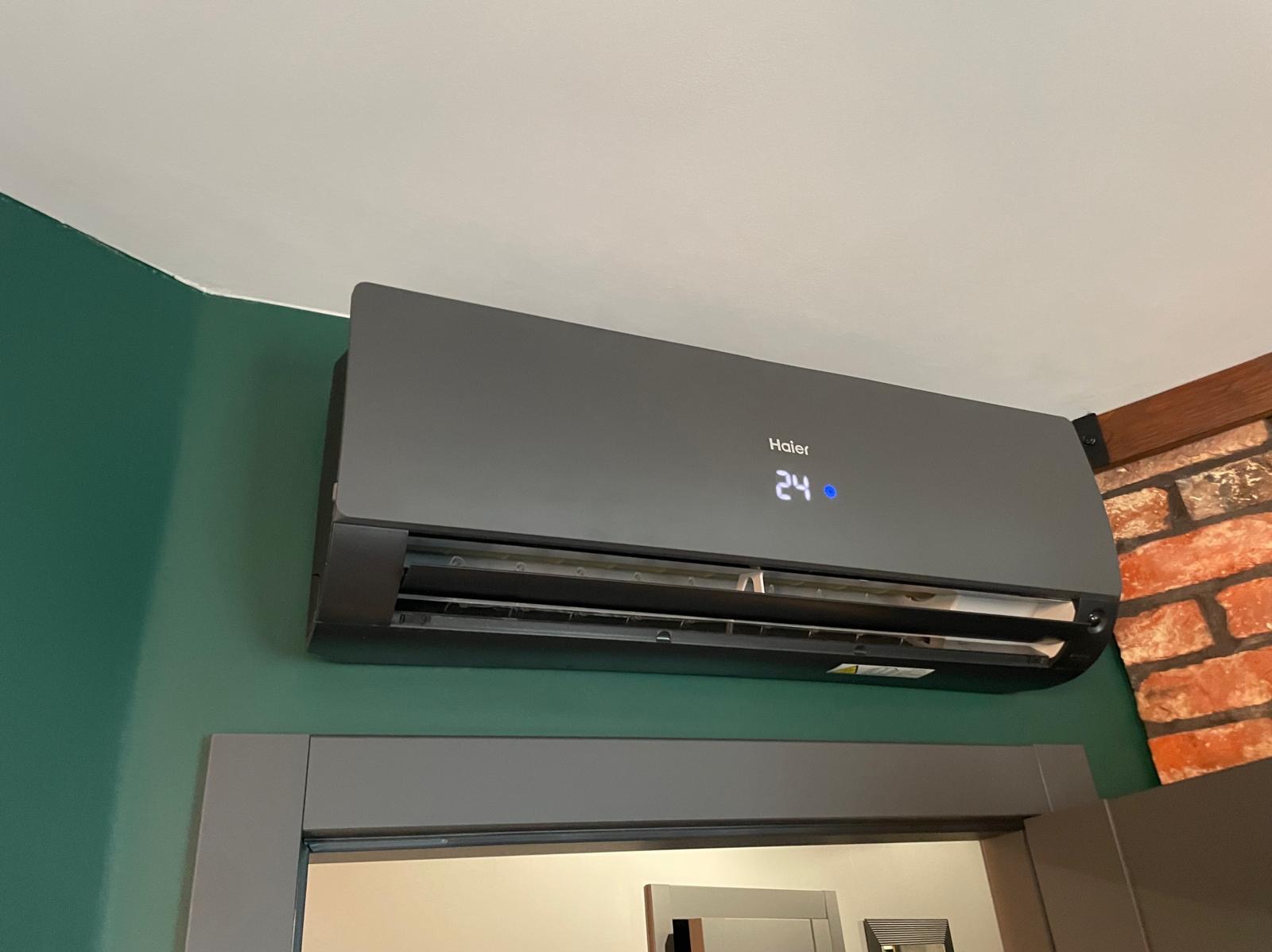 Grafitowy klimatyzator Haier zamontowany na zielonej ścianie, pod sufitem, obok ceglanej ściany i szarej futryny drzwi. Na wyświetlaczu widoczna temperatura 24 stopnie.