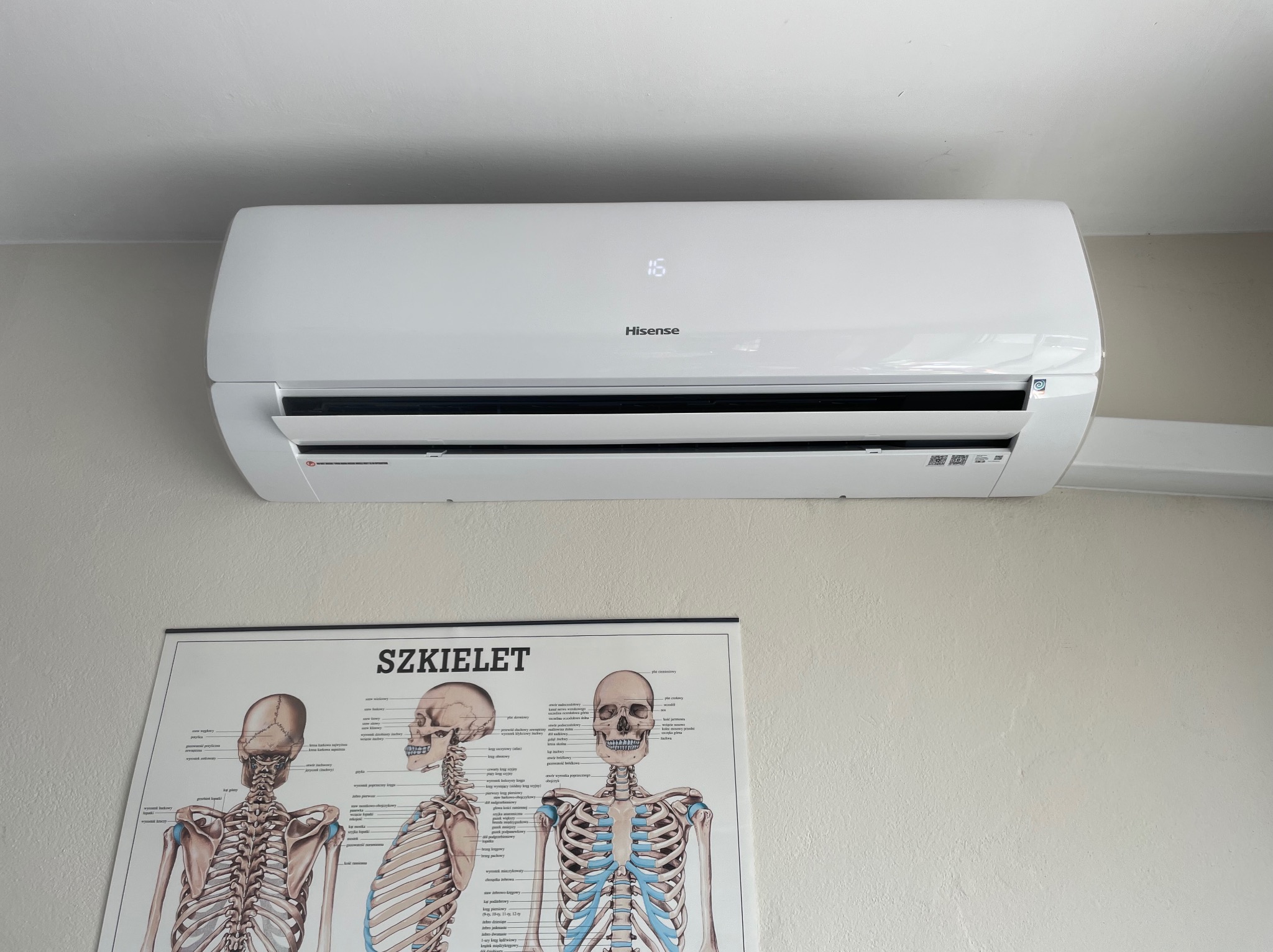 Biały klimatyzator marki Hisense zamontowany na ścianie w pomieszczeniu, widoczny wyświetlacz z temperaturą 16 stopni, poniżej plakat edukacyjny ze schematem szkieletu człowieka.