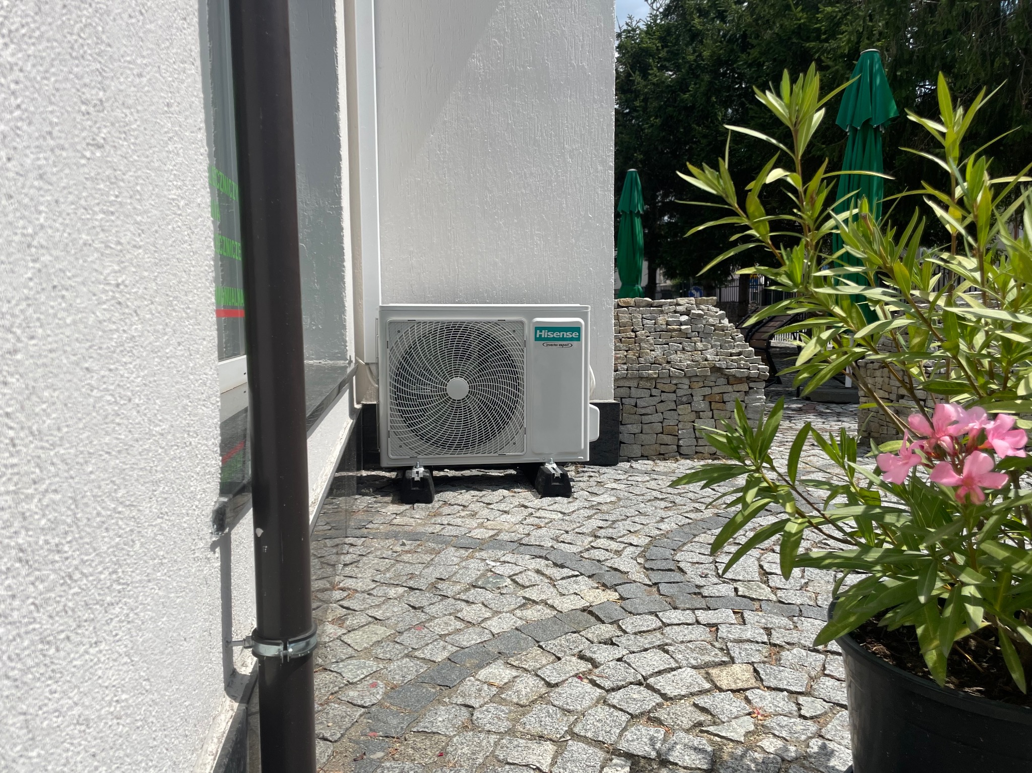 Biała jednostka zewnętrzna klimatyzacji Hisense zamontowana na czarnych wspornikach przy białej ścianie budynku, na tle brukowanej nawierzchni i roślinności.