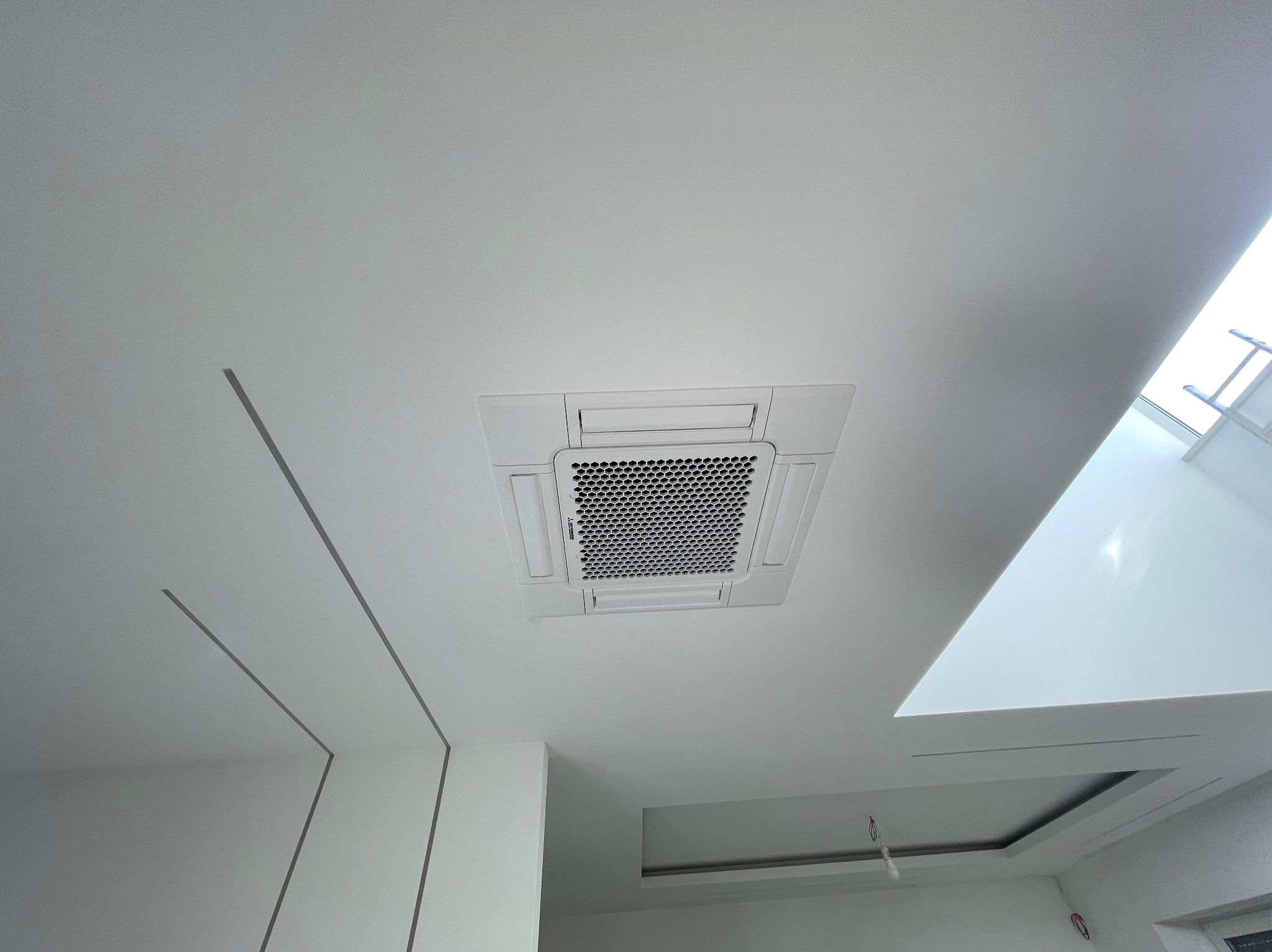 Kasetonowa jednostka klimatyzacyjna zamontowana w białym suficie podwieszanym, widok z dołu, widoczne minimalistyczne linie dekoracyjne na ścianie.