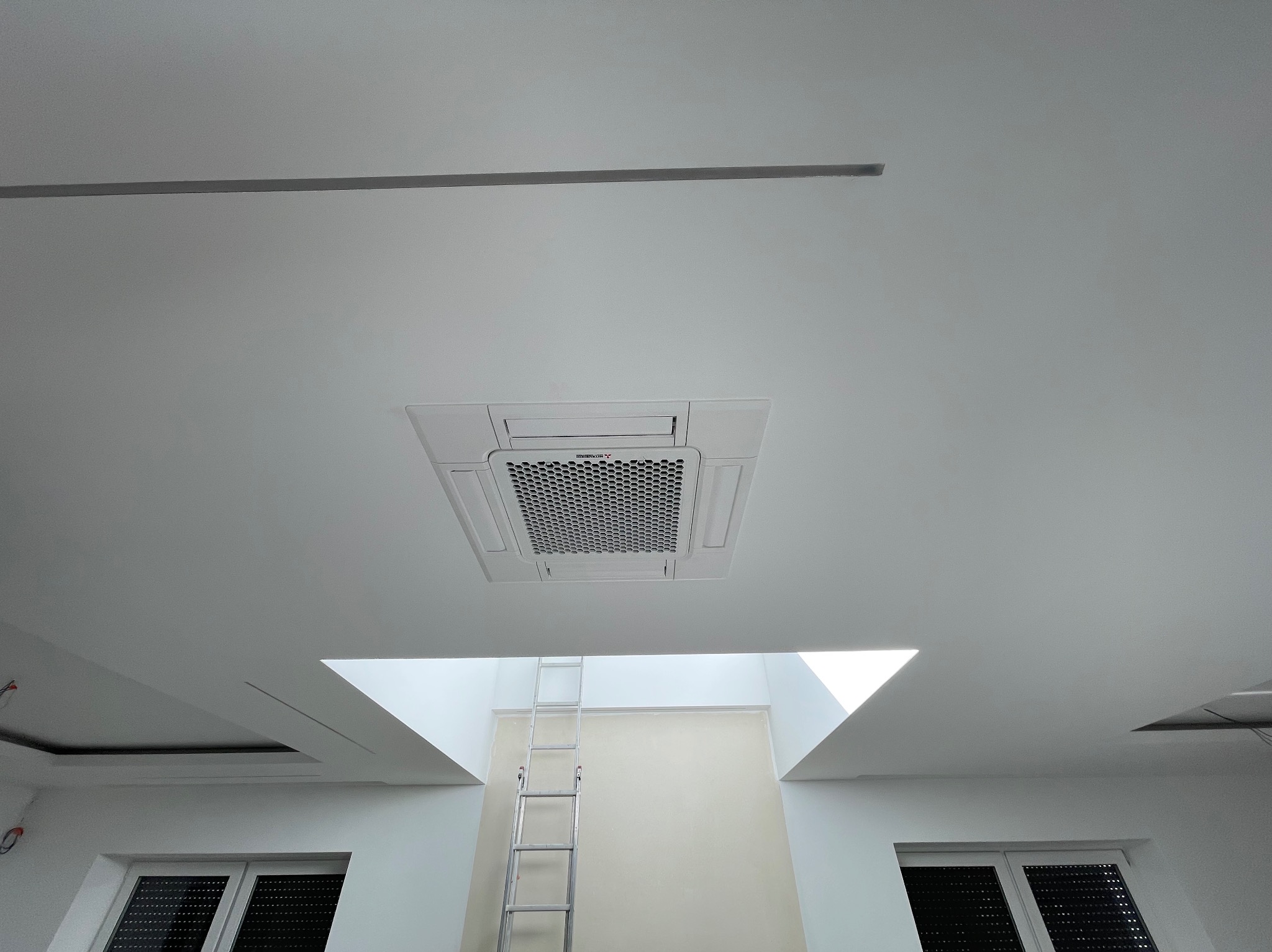 Kasetonowa jednostka wewnętrzna systemu klimatyzacji zamontowana w suficie podwieszanym, widok z dołu, w tle drabina oparta o ścianę z oknem dachowym.