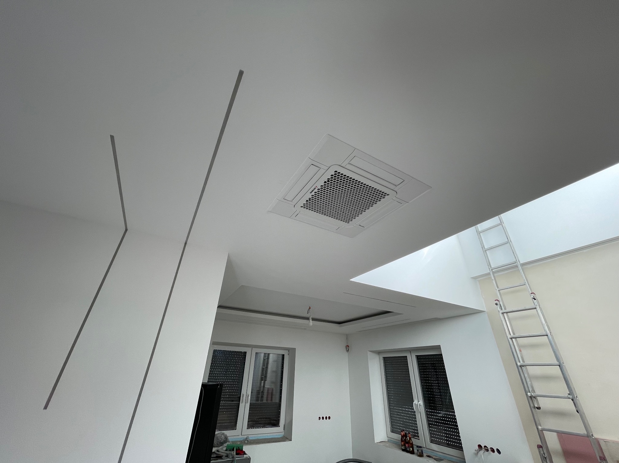 Kasetonowa jednostka klimatyzacyjna zamontowana w suficie podwieszanym w jasnym pomieszczeniu, widoczna drabina oparta o ścianę, fragmenty dekoracyjnych listew sufitowych.