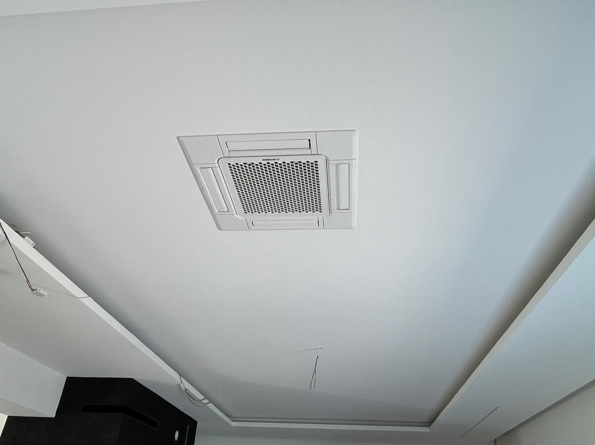 Biała kaseta sufitowa klimatyzatora zamontowana na białym suficie podwieszanym z widocznymi elementami oświetlenia i instalacji elektrycznej.