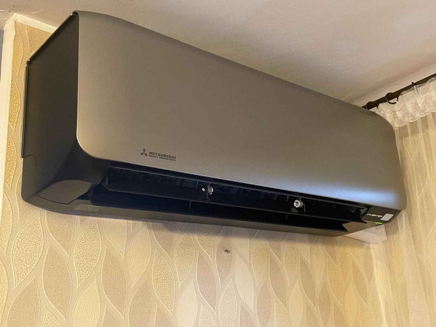 Zamontowany, srebrno-czarny klimatyzator Mitsubishi na ścianie z wzorzystą tapetą. Widoczny fragment firanki. Urządzenie gotowe do użycia.