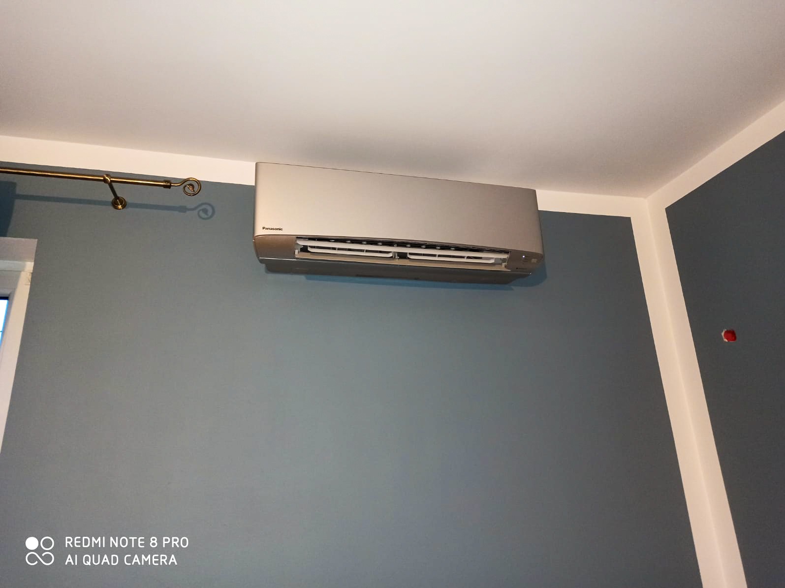 Zamontowany klimatyzator Panasonic na szarej ścianie w pomieszczeniu, widoczna listwa karnisza i narożnik ściany z białą lamperią. Urządzenie w kolorze srebrnym.