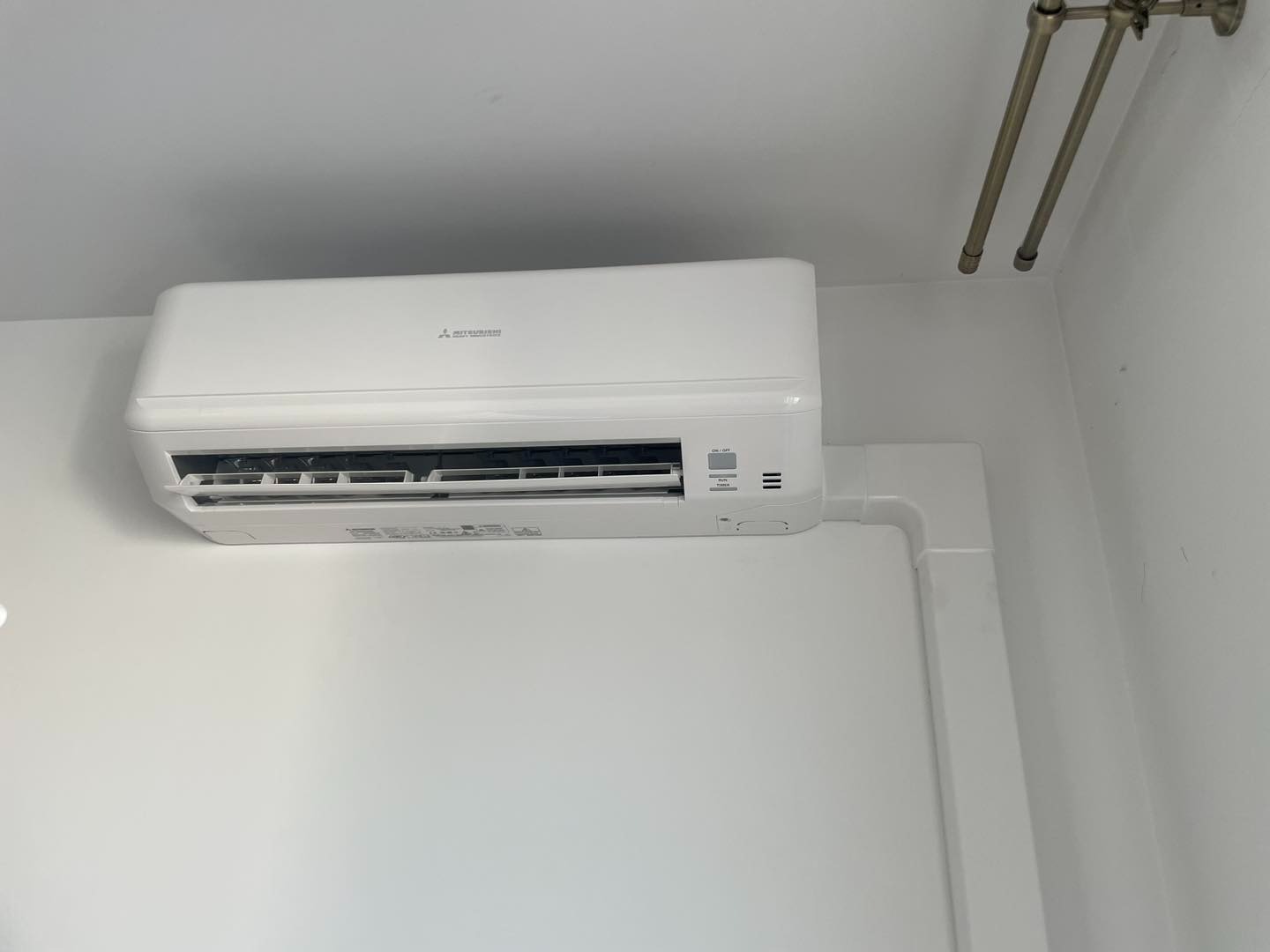 Biały klimatyzator Mitsubishi, zamontowany na ścianie, z widocznymi przyciskami sterowania i częściowo otwartą klapą nawiewu. U góry widoczne rurki.