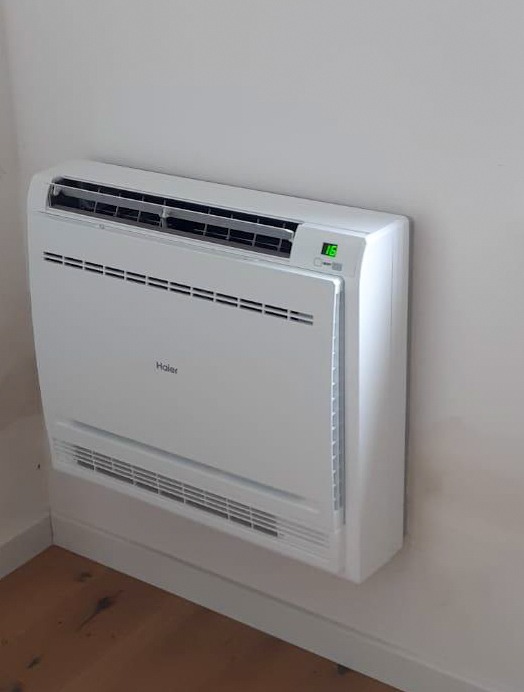 Biały, przypodłogowy klimatyzator Haier z wyświetlaczem temperatury (16°C) zamontowany przy ścianie na tle jasnego wnętrza z drewnianą podłogą.