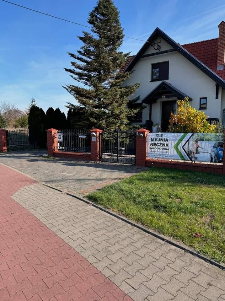 Ogrodzenie posesji z reklamą myjni ręcznej samochodowej na tle domu z dużym drzewem iglastym.